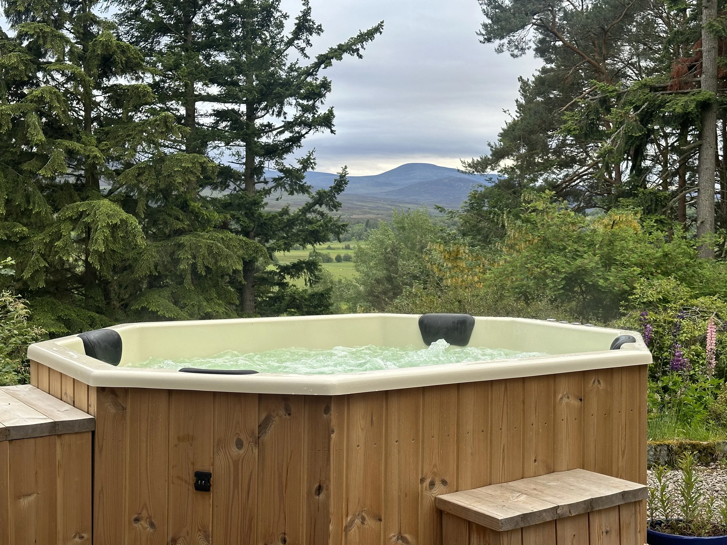 Hot tub at Ardchoile, Kingussie with water jet streaming, surrounded by wooden siding, located outdoors among lush trees with a mountain in the background.