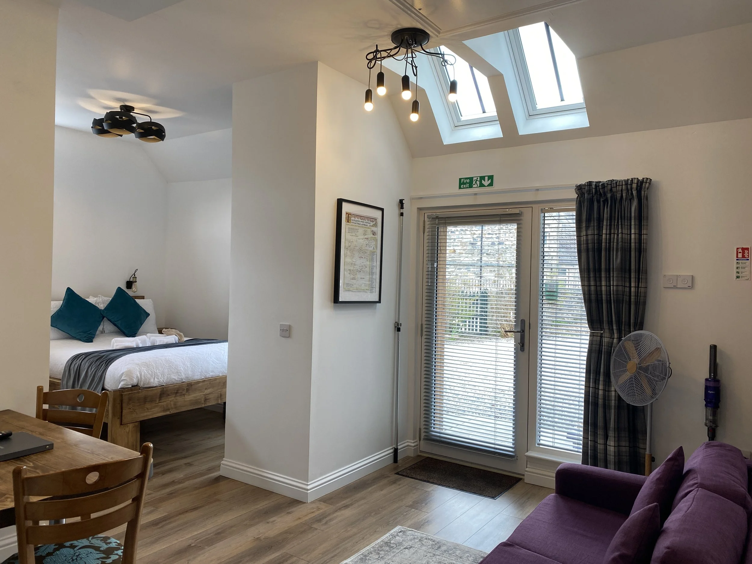 Interior of a bright, modern apartment featuring a bedroom with a wooden bed, teal pillows, and white bedding visible in a separate area, a dining table with chairs, a purple sofa, a glass door with blinds and plaid curtains, skylights, and contemporary ceiling lights.