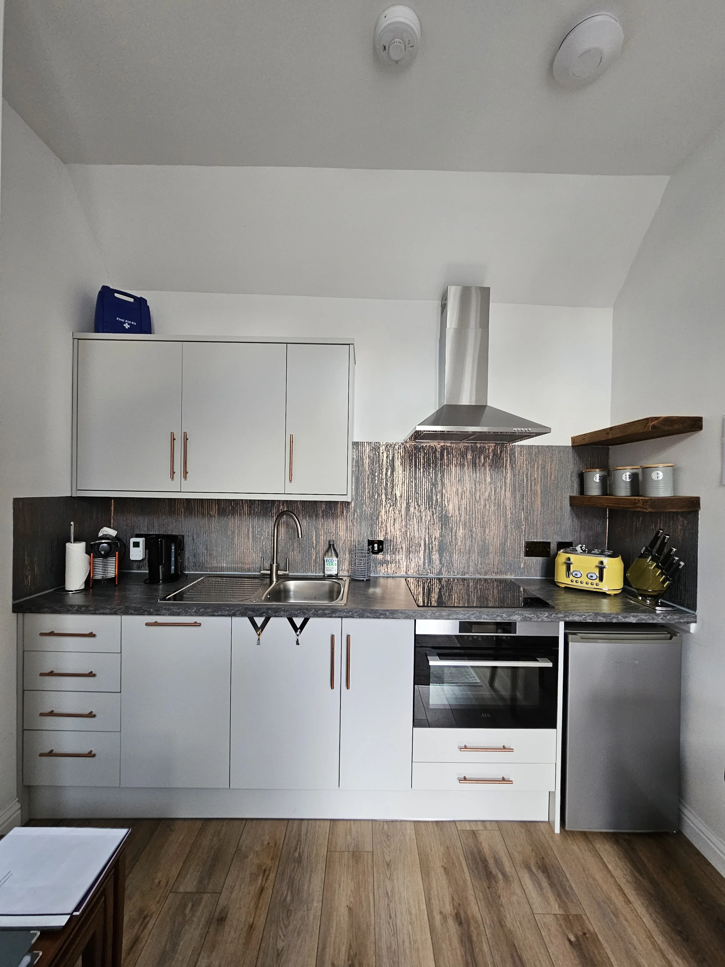 Modern kitchen with white cabinets, copper handles, a stainless steel sink, a stove, and a small refrigerator. Black and yellow appliances on the counter, open wooden shelves with storage jars, wood flooring, and a copper backsplash.