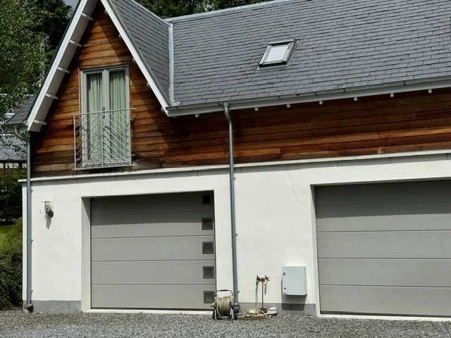 Modern house with two garage doors, wooden and white exterior, gravel driveway, benches, and potted plants, surrounded by trees.