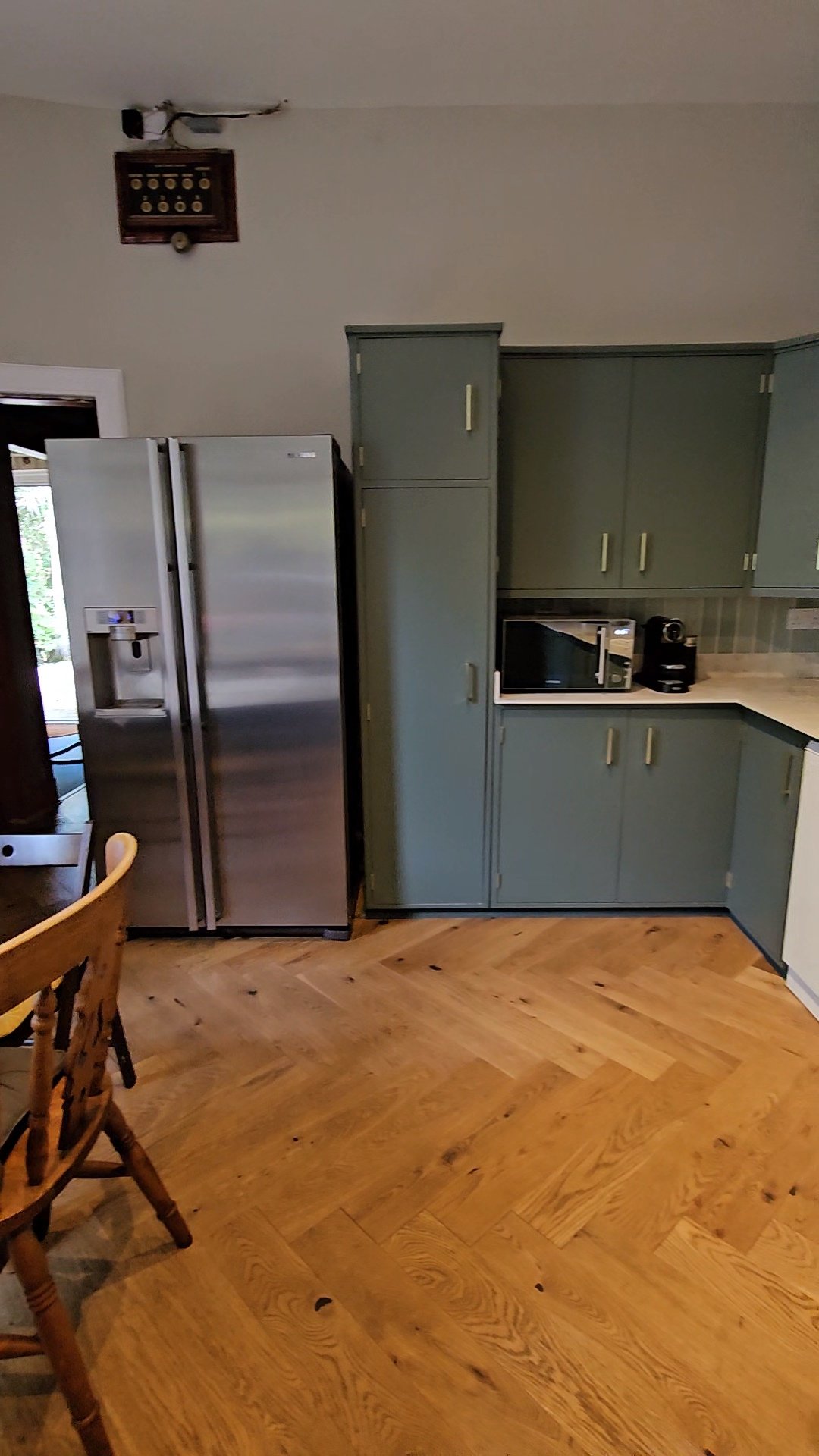 A kitchen with a stainless steel side-by-side refrigerator, green cabinets, a microwave, and a coffee maker on the counter. There is wood flooring and a wooden chair partially visible in the foreground. An electrical box with exposed wires is mounted on the wall near the ceiling.