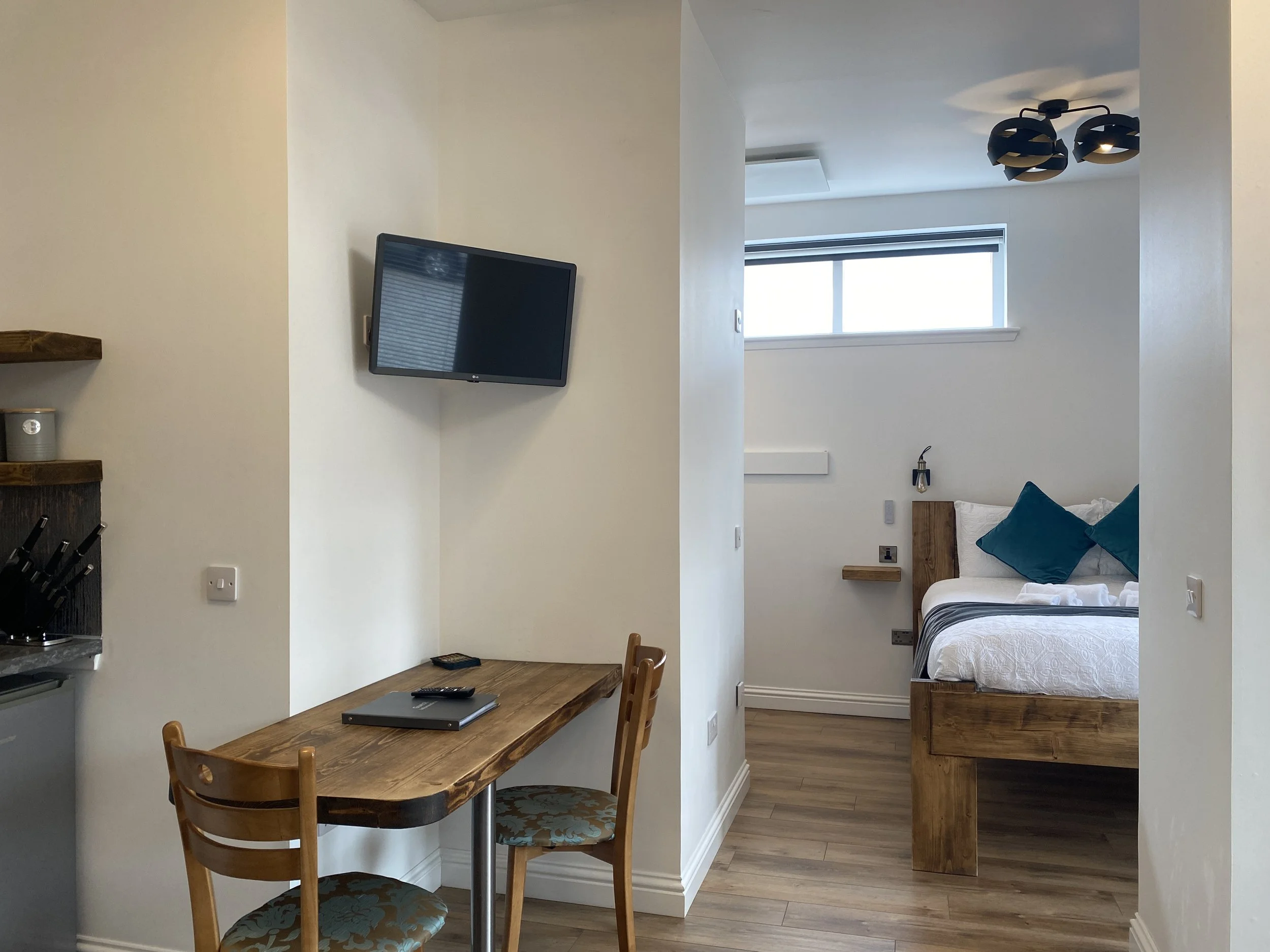 A small hotel room with a breakfast bar, two chairs, and a wall-mounted TV. In the background, a bed with teal pillows, bedside lights, and a window with a blind.