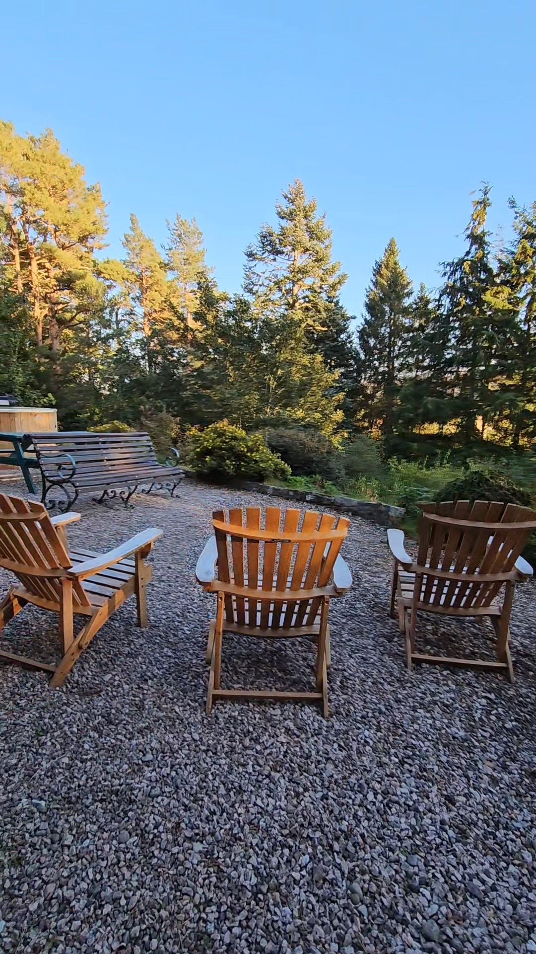 Outdoor seating area with wooden chairs and benches on gravel, surrounded by trees under a clear blue sky.
