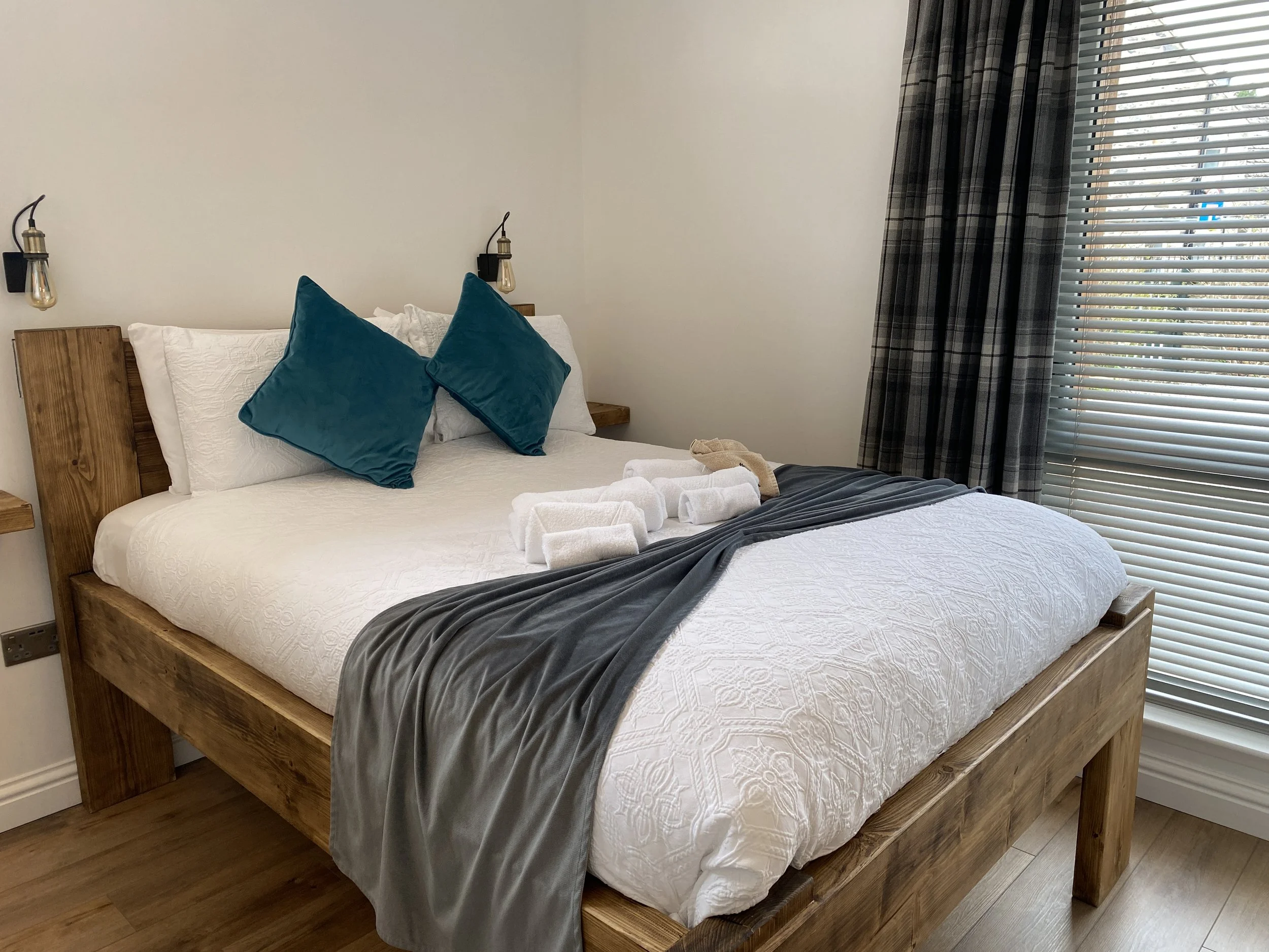 A neatly made bed with white bedding, two dark teal pillows, a gray throw blanket, and folded white towels on top. The bed has a wooden frame and headboard, with black wall-mounted lamps on either side. There is a window with partially open blinds and plaid curtains on the right side of the room.