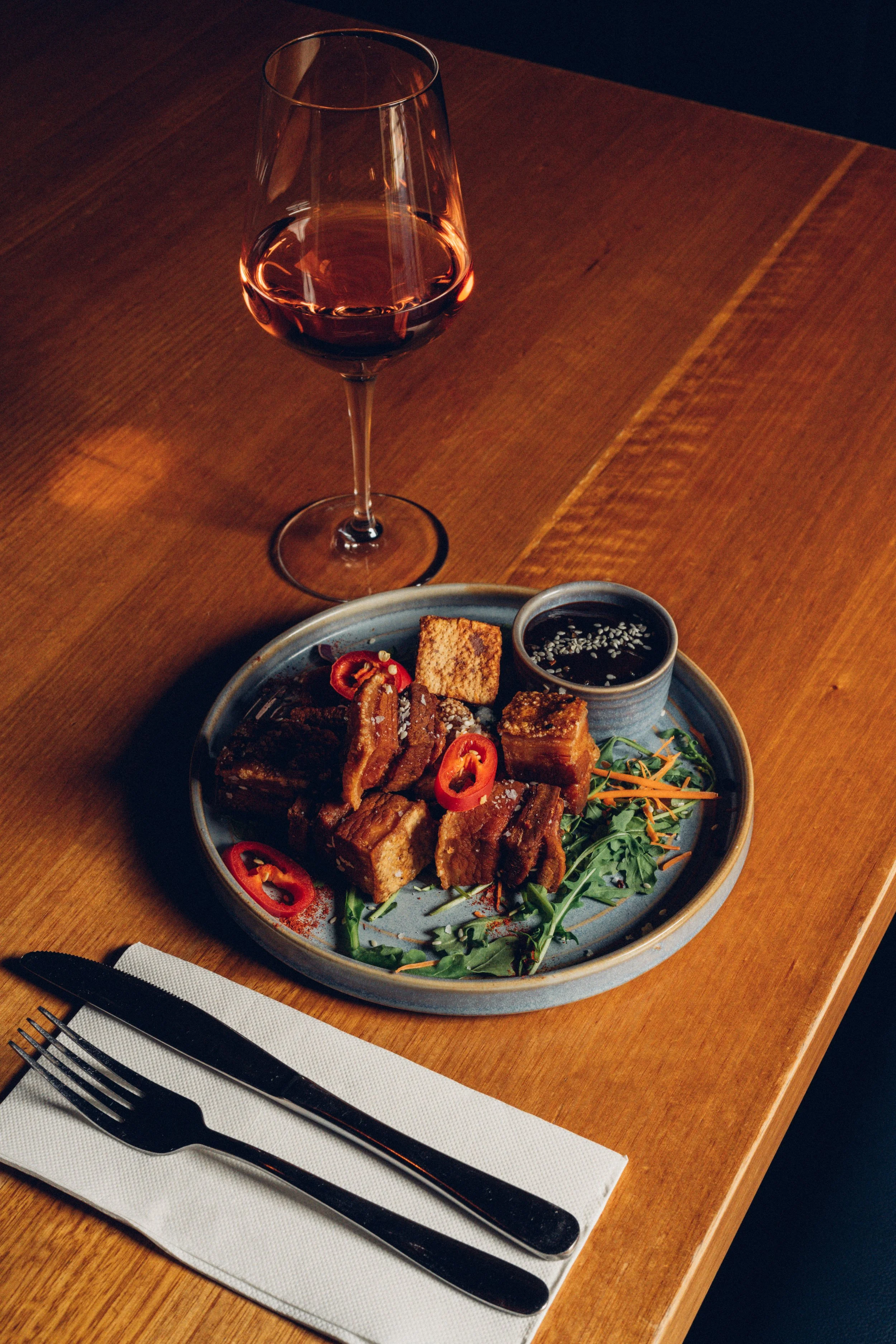 A glass of red wine on a wooden table, a plate with assorted fried foods, sliced red peppers, a small bowl of dipping sauce, a fork, knife, and napkin.