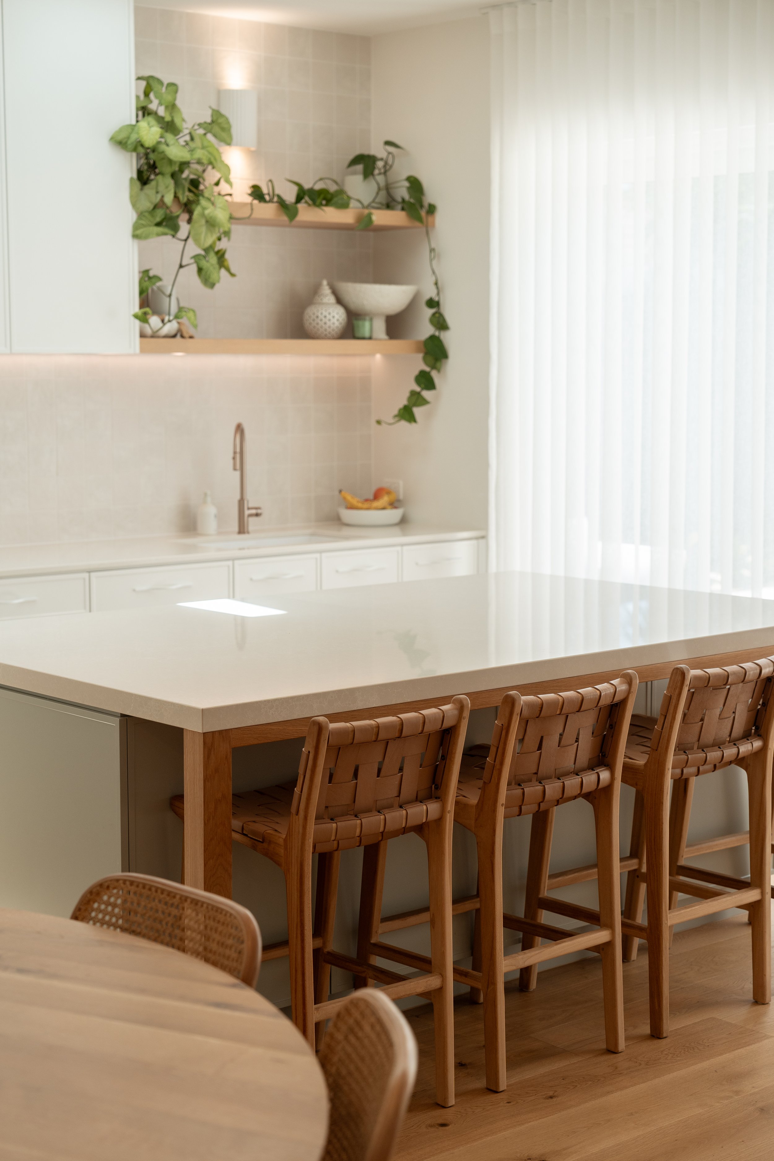 Modern kitchen with a white countertop, wooden bar stools, and open shelves decorated with plants and ceramic bowls, illuminated by natural light from a large window with sheer curtains.