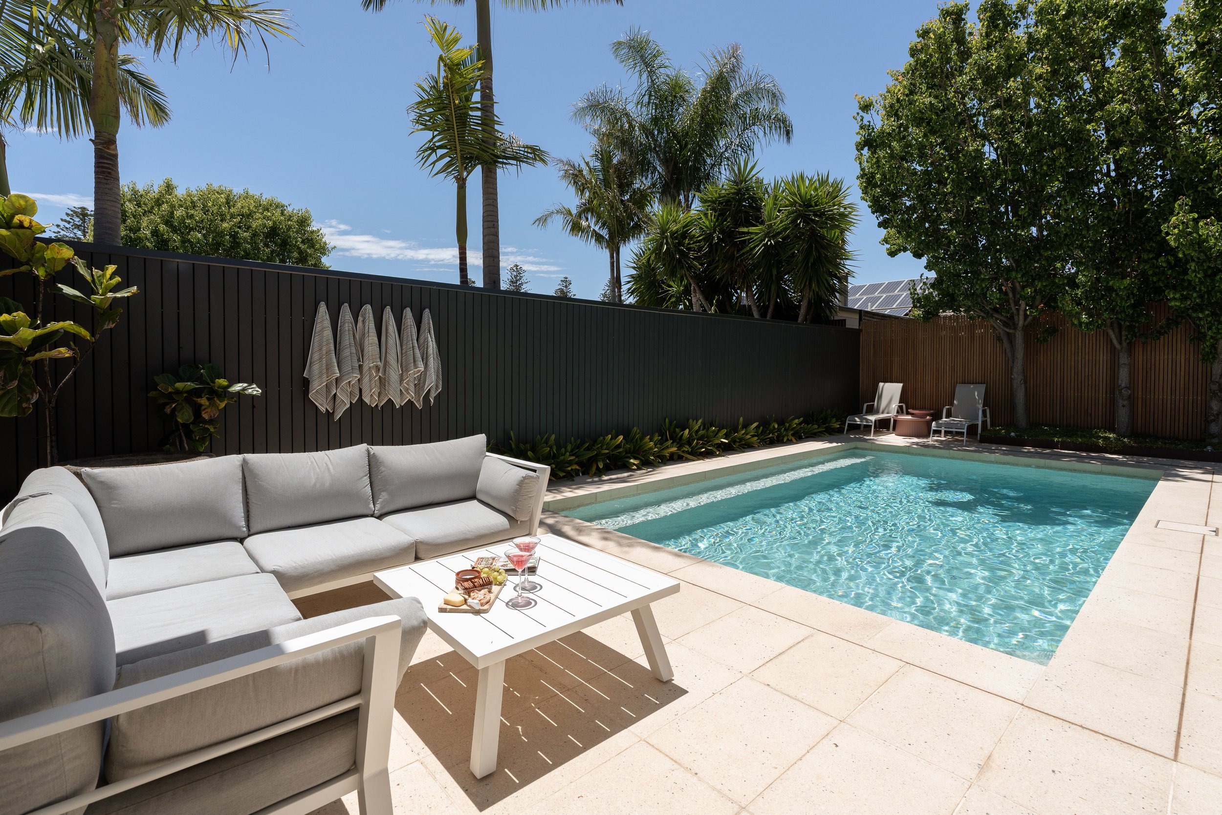 Backyard swimming pool with patio furniture, umbrellas, and lush greenery under a clear blue sky.