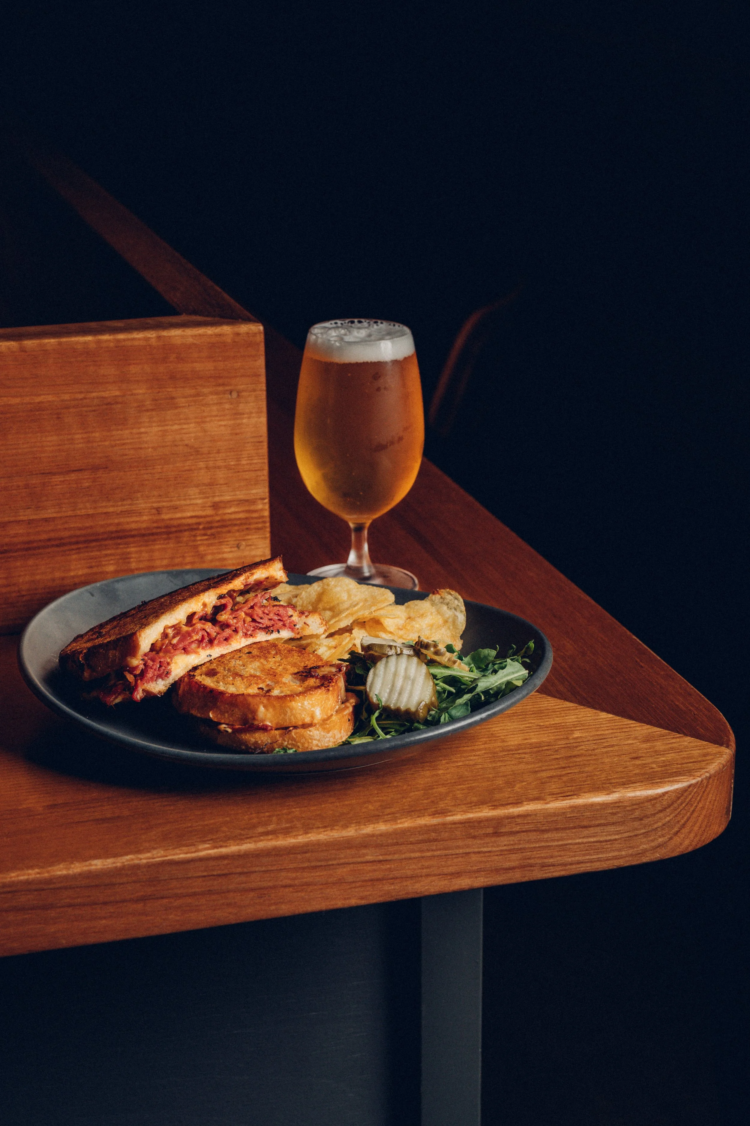 A plate of club sandwich with chips and greens, a glass of beer on a wooden table in a dimly lit setting.
