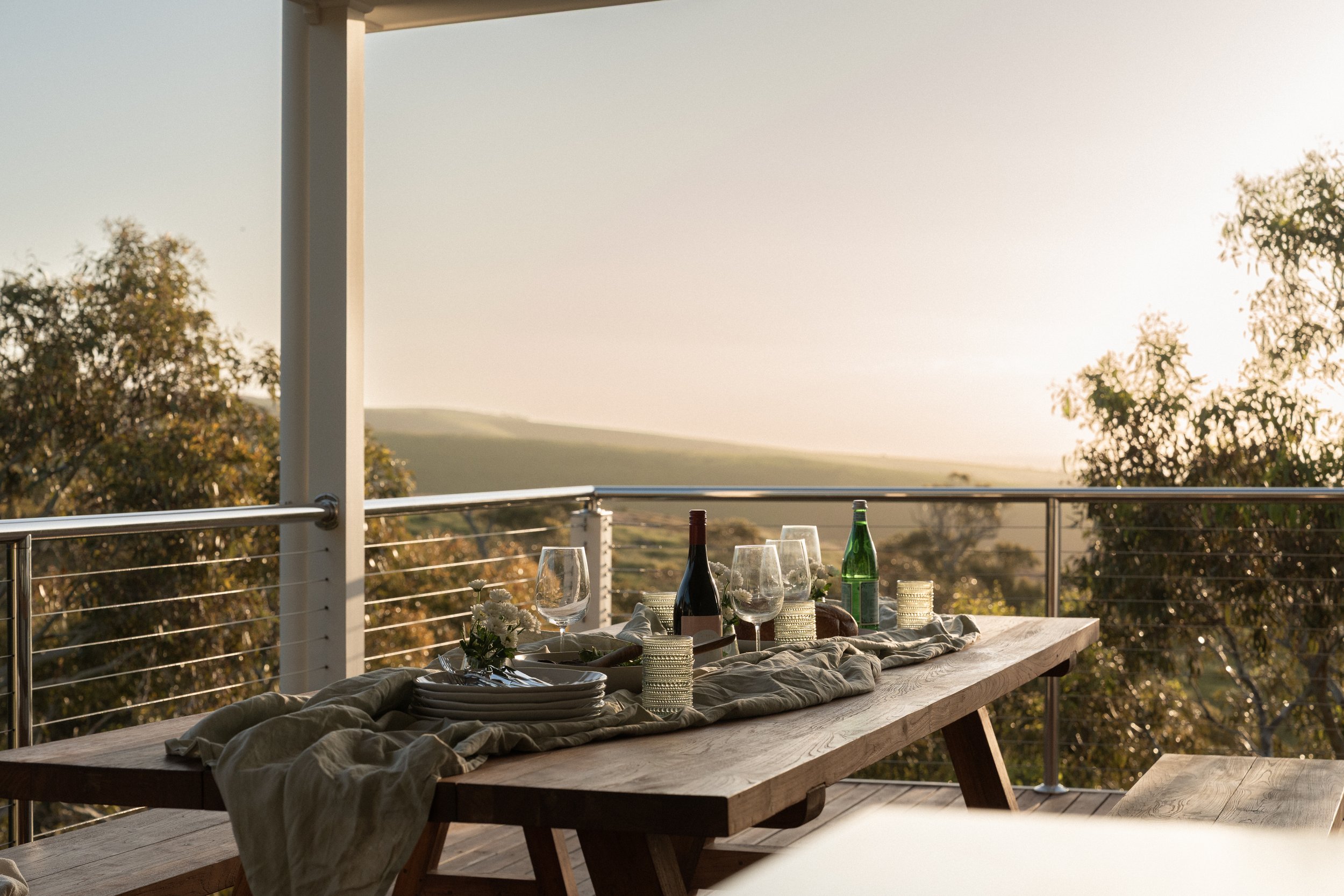 A wooden outdoor table set with wine glasses, plates, candles, and a centerpiece of white flowers, overlooking a landscape with trees and hills during sunset.