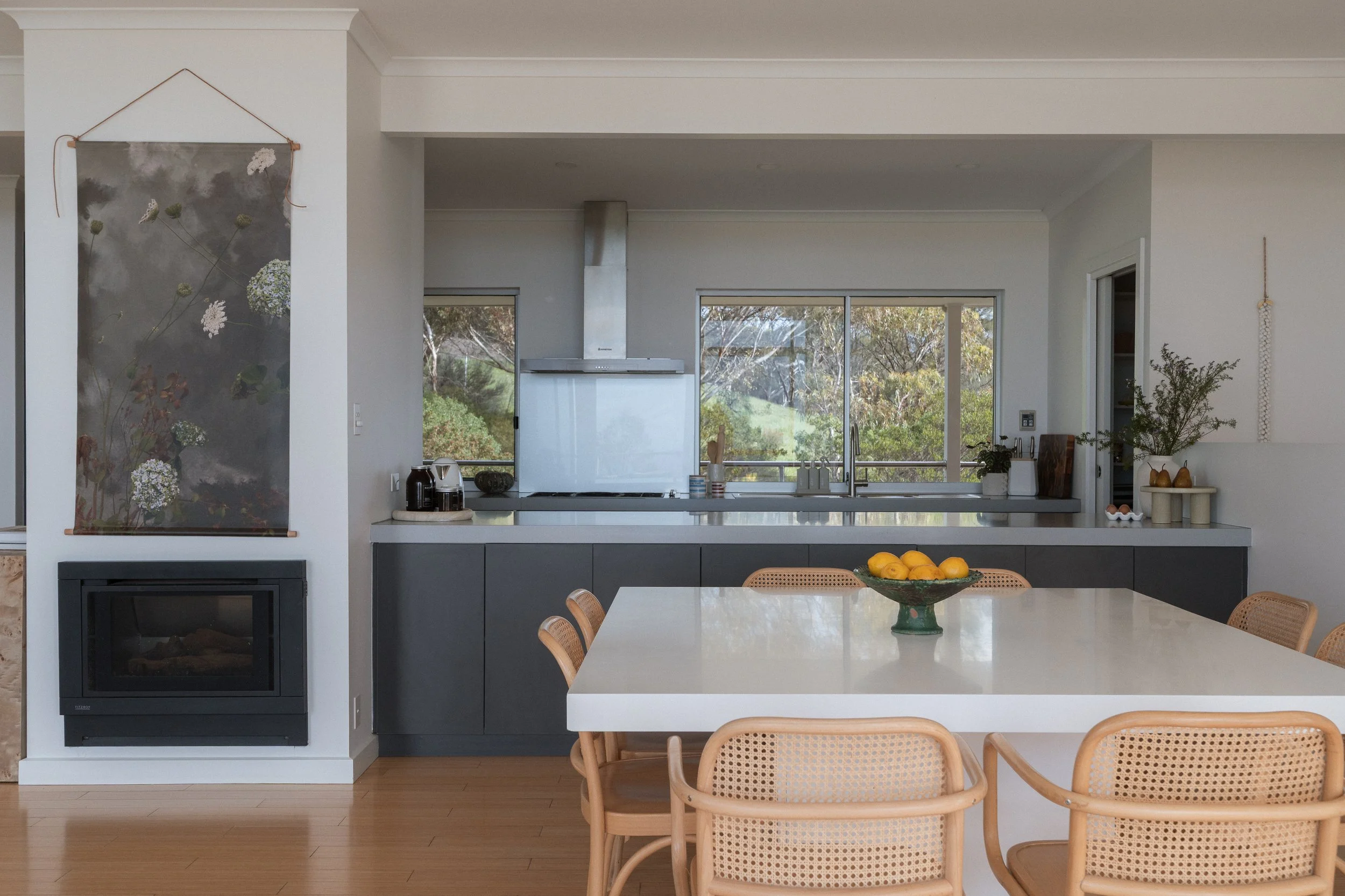 Modern kitchen interior with white countertops, large windows overlooking greenery, a dark gray lower cabinet, and a white table with wicker chairs. Decor includes a bowl of lemons and potted plants.