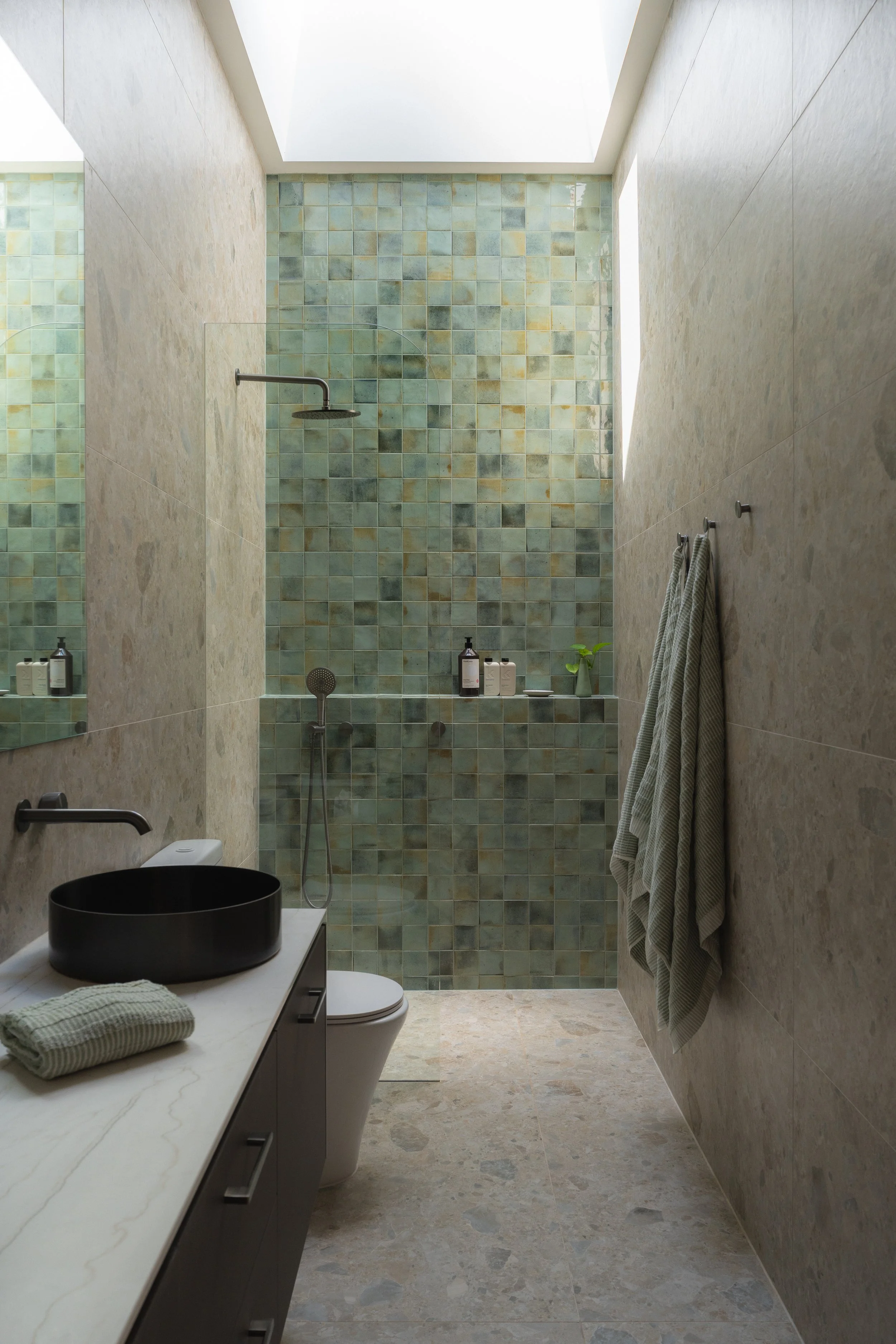Modern bathroom featuring a walk-in shower with green mosaic tiles, a black vessel sink on a white marble countertop, a wall-mounted faucet, a toilet, and striped towels hanging on hooks.