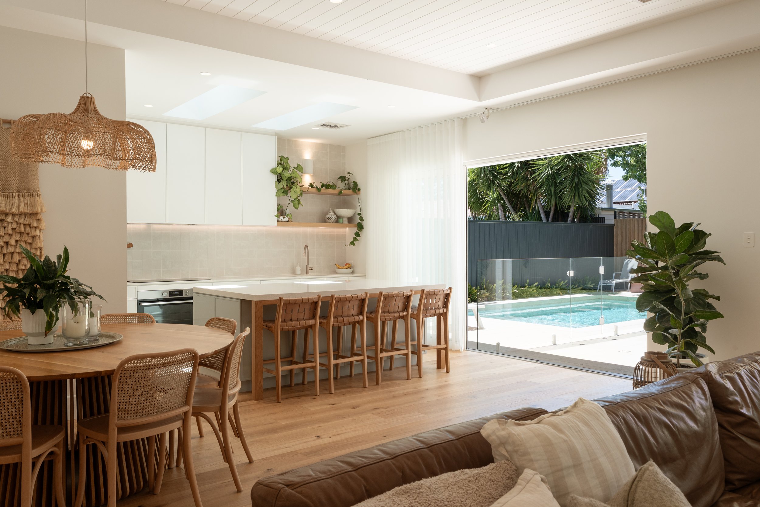 Open-concept living space with a view of a backyard pool through large glass sliding doors, featuring a kitchen with a white island and bar stools, a round dining table with chairs, and indoor plants.