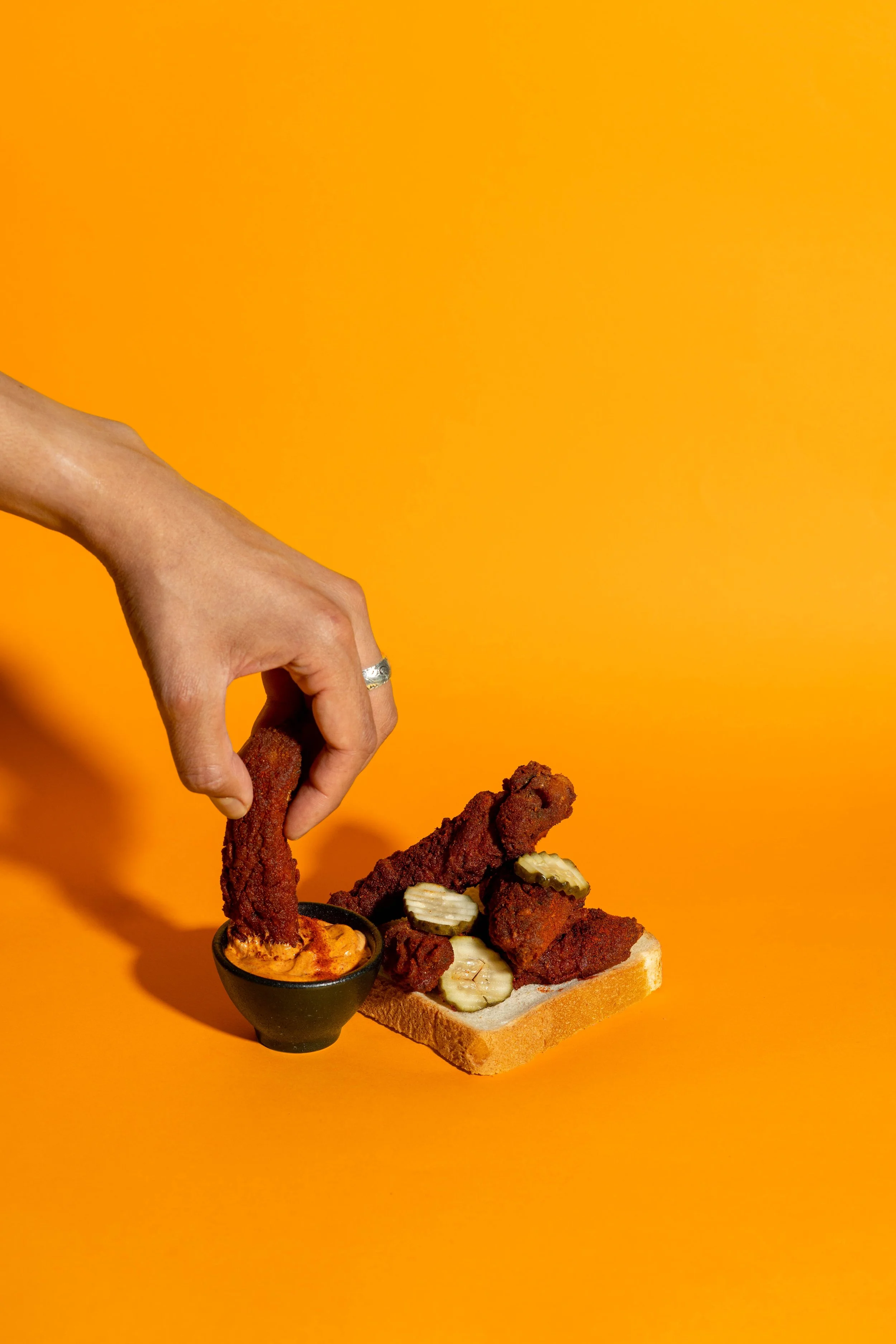 A hand with a ring drops fried chicken strips into a small cup of orange dipping sauce. There is a pizza slice topped with pickles and fried chicken strips on a piece of bread. The background is bright orange.