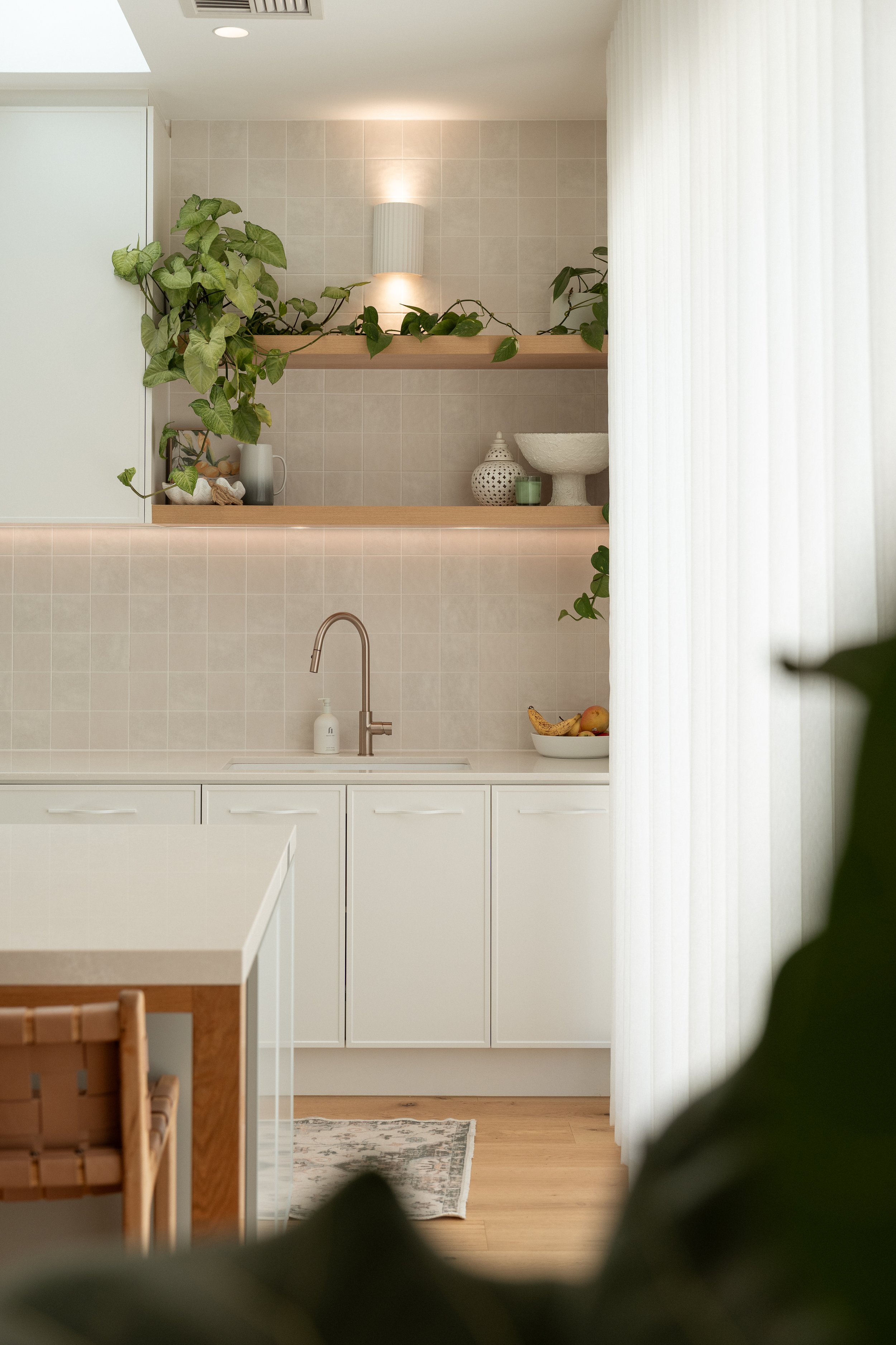 A minimalist kitchen with white cabinets, a beige tiled backsplash, and a stainless steel faucet on the sink. There are wooden shelves above the sink decorated with plants, ceramic vases, and a stone bowl. To the right, a bowl of fruit is visible, an