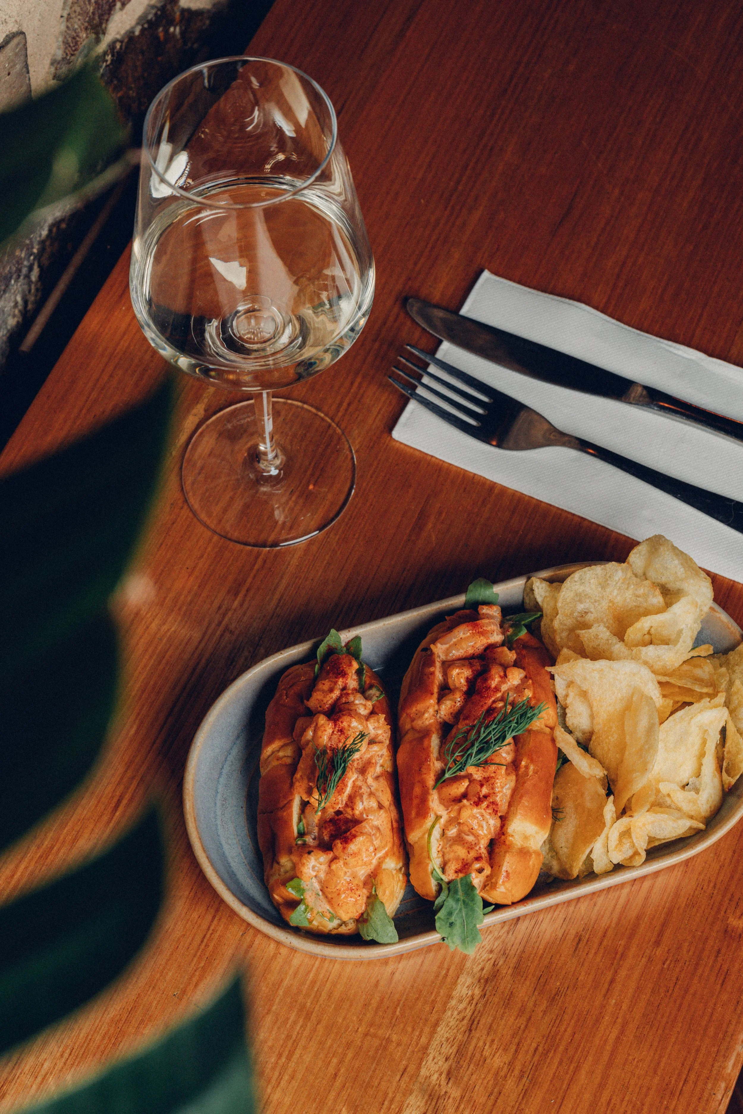 A glass of water on a wooden table next to a white napkin with a knife and fork on top, and a plate of hot dog buns with baked chicken, potatoes, and potato chips garnished with herbs.