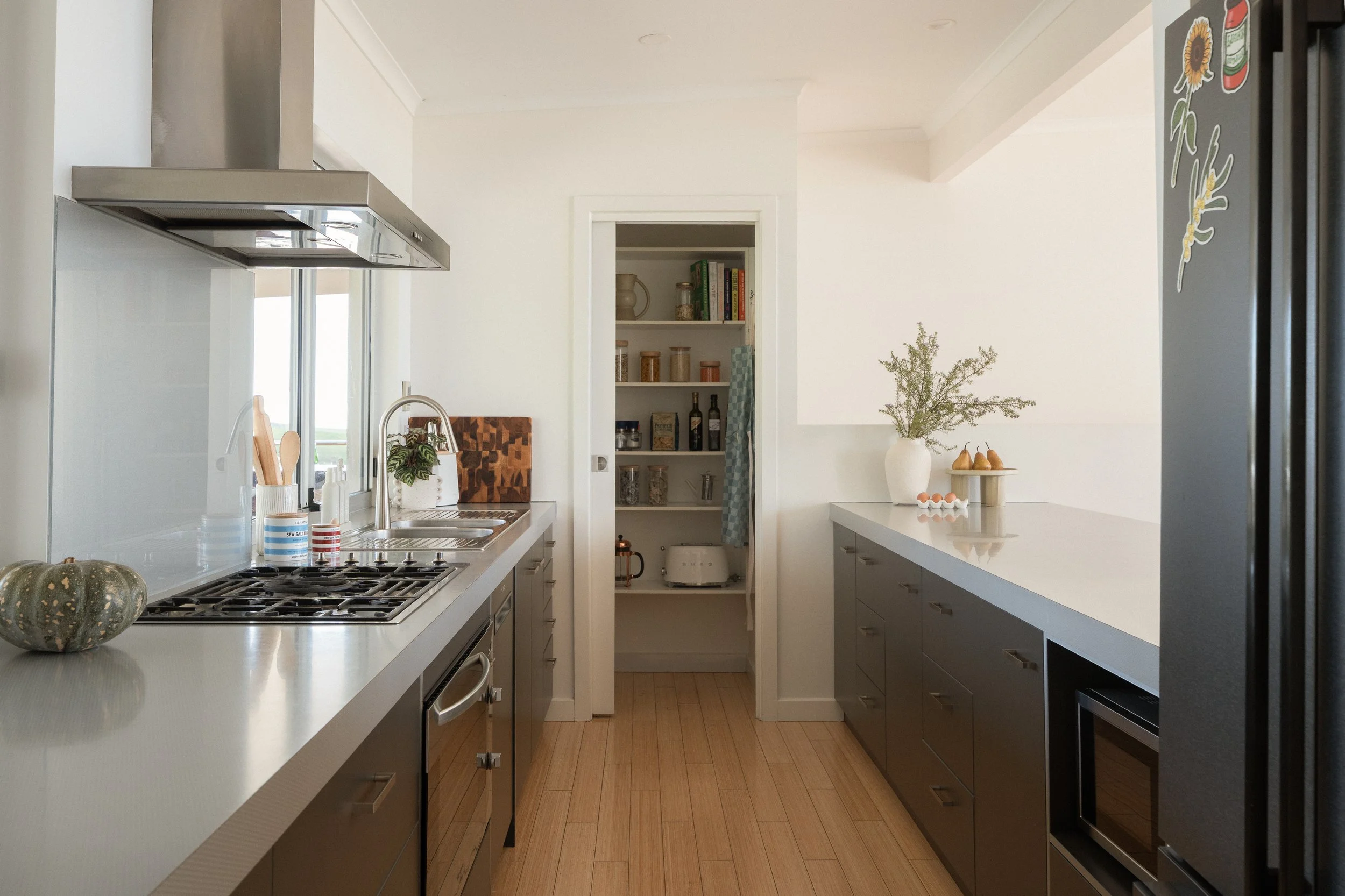 A modern kitchen with white countertops, dark lower cabinets, and stainless steel appliances. There is a pumpkin on the counter, a potted plant, and a window with a view outside. An open pantry with shelves containing jars, bottles, and books is visi
