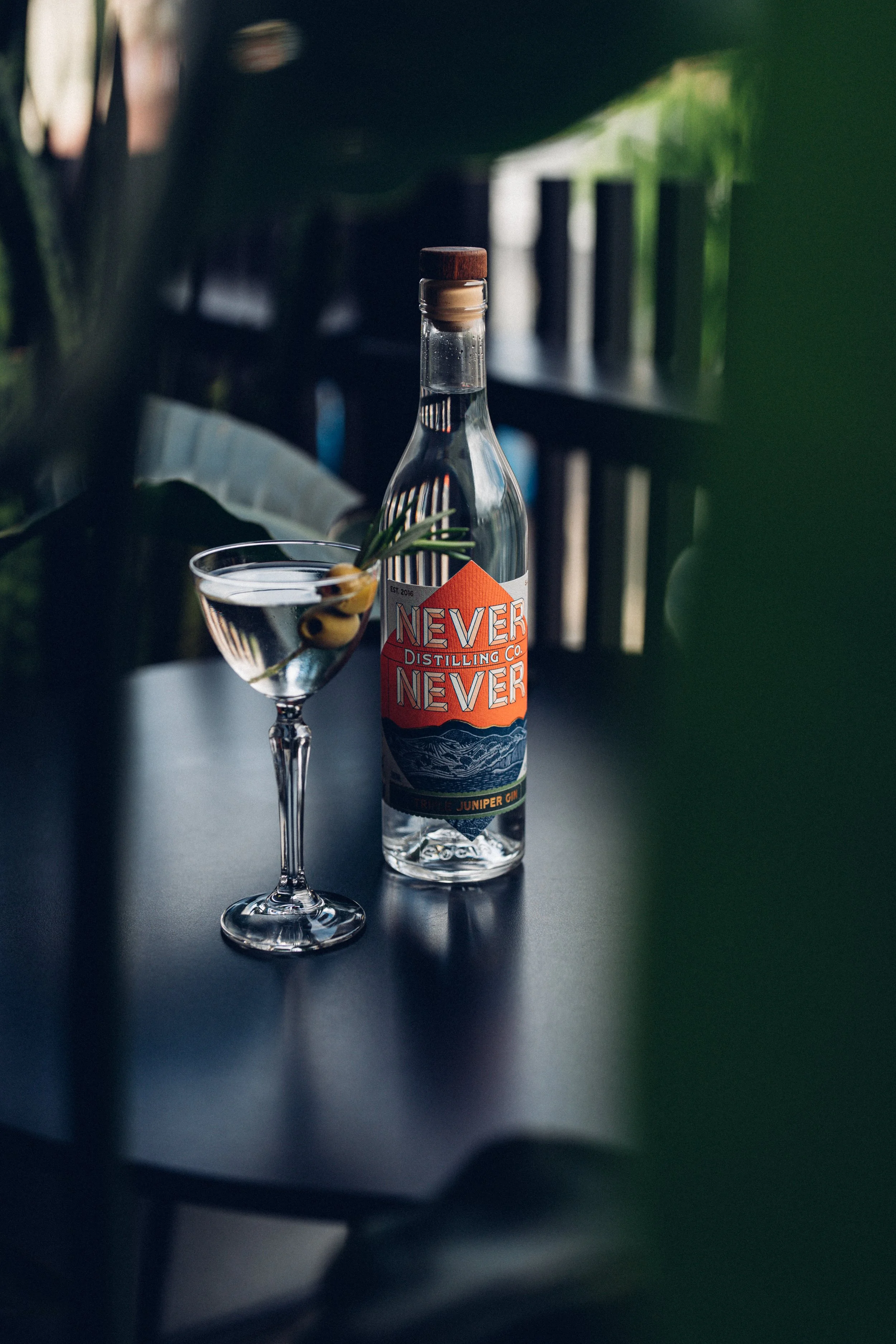 A bottle of Never Never Distilling Co. juniper-infused gin and a cocktail garnished with olives and herbs on a table.