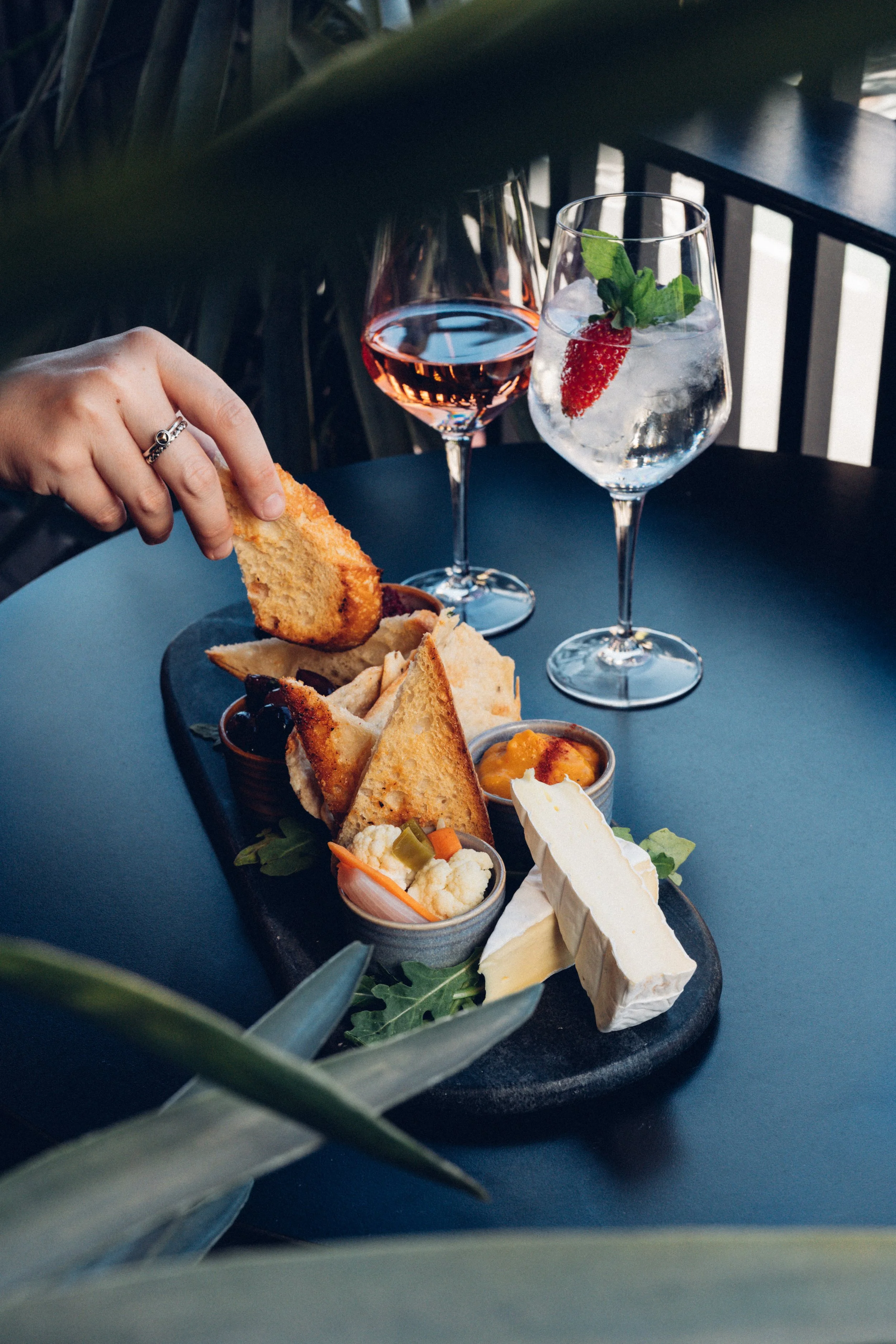 A black serving platter filled with a variety of cheeses, crackers, and small bowls of vegetables and sauces, with two glasses of rosé wine and a glass of water with a strawberry and mint.