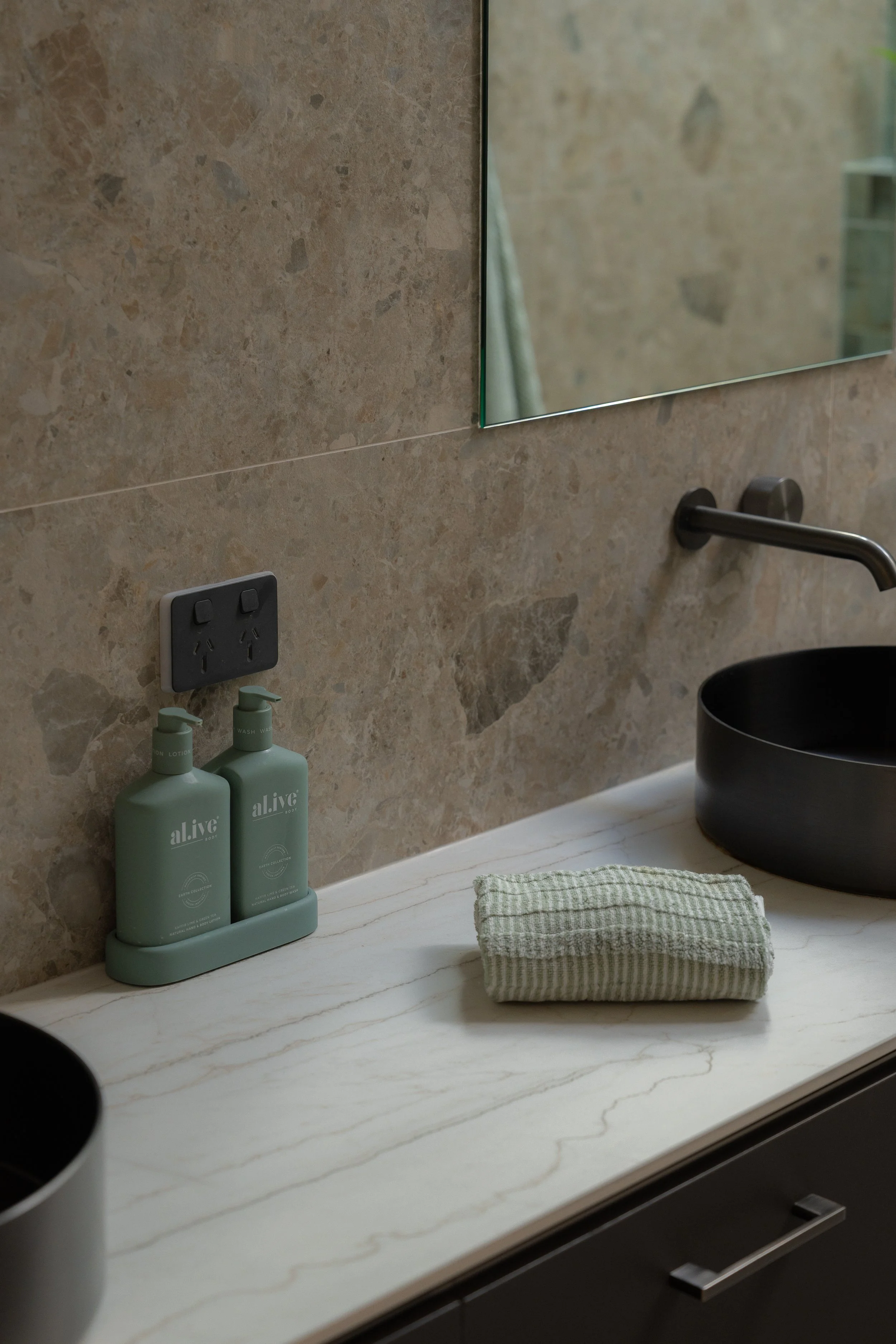 A bathroom countertop with a black round sink, a folded green and white striped towel, two green bottles labeled 'alive' for lotion and wash, a mirror, a wall-mounted faucet, and a power outlet with two roles of stamps or labels.