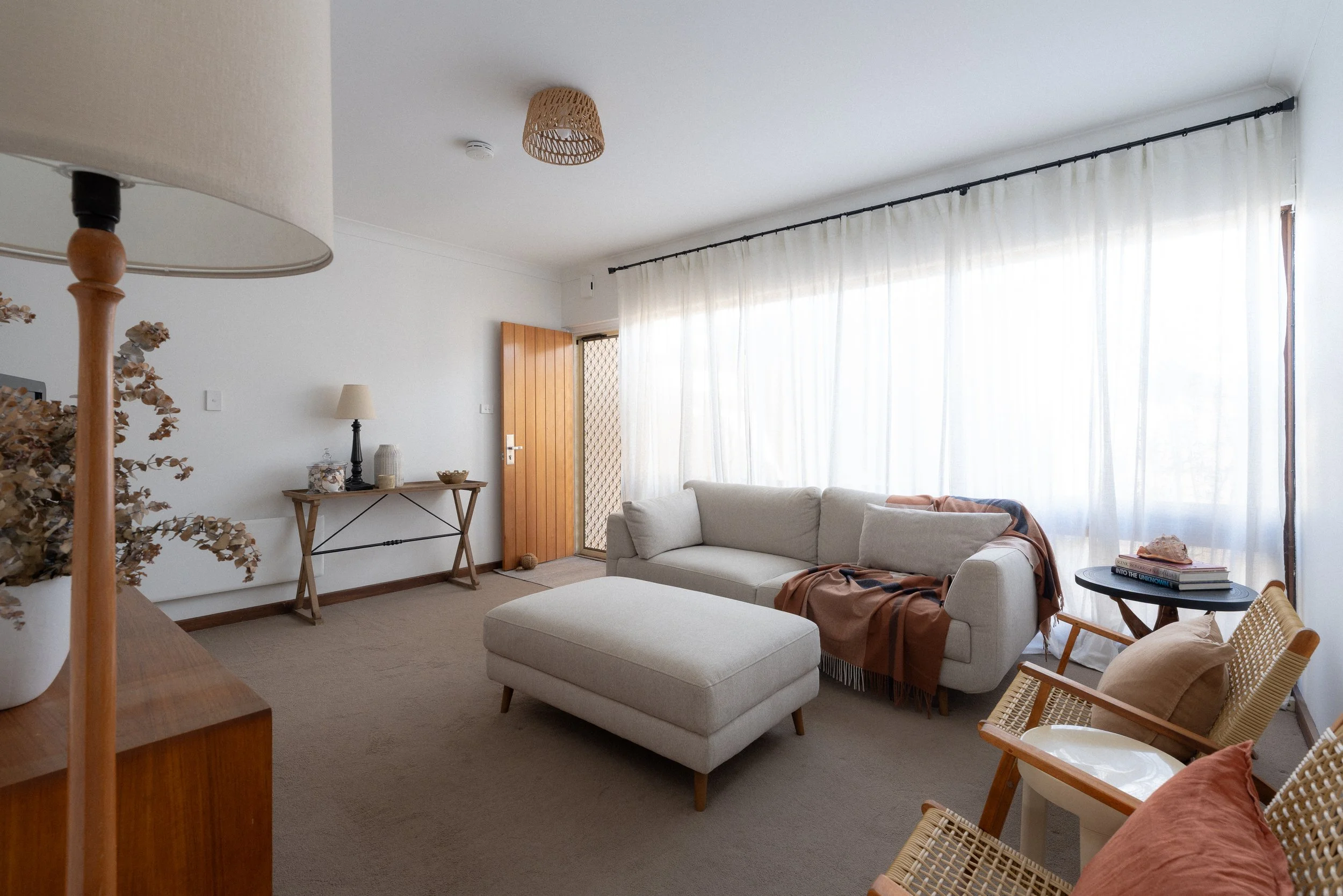 Living room with a beige sofa, a wooden door, a side table with books, a table lamp, and sheer curtains allowing sunlight.