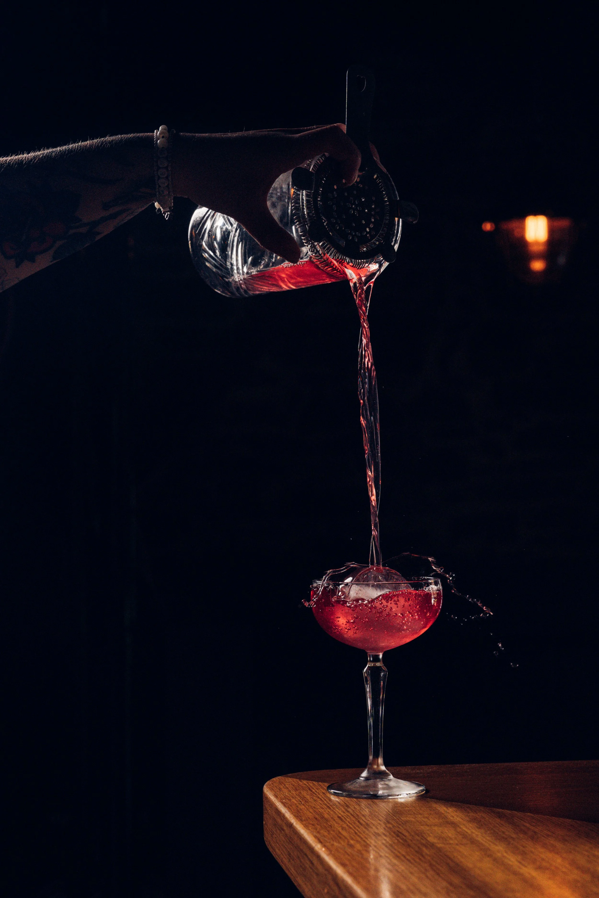 A hand with tattoos pouring red liquid from a plastic water bottle into a cocktail glass filled with red liquid, against a dark background.