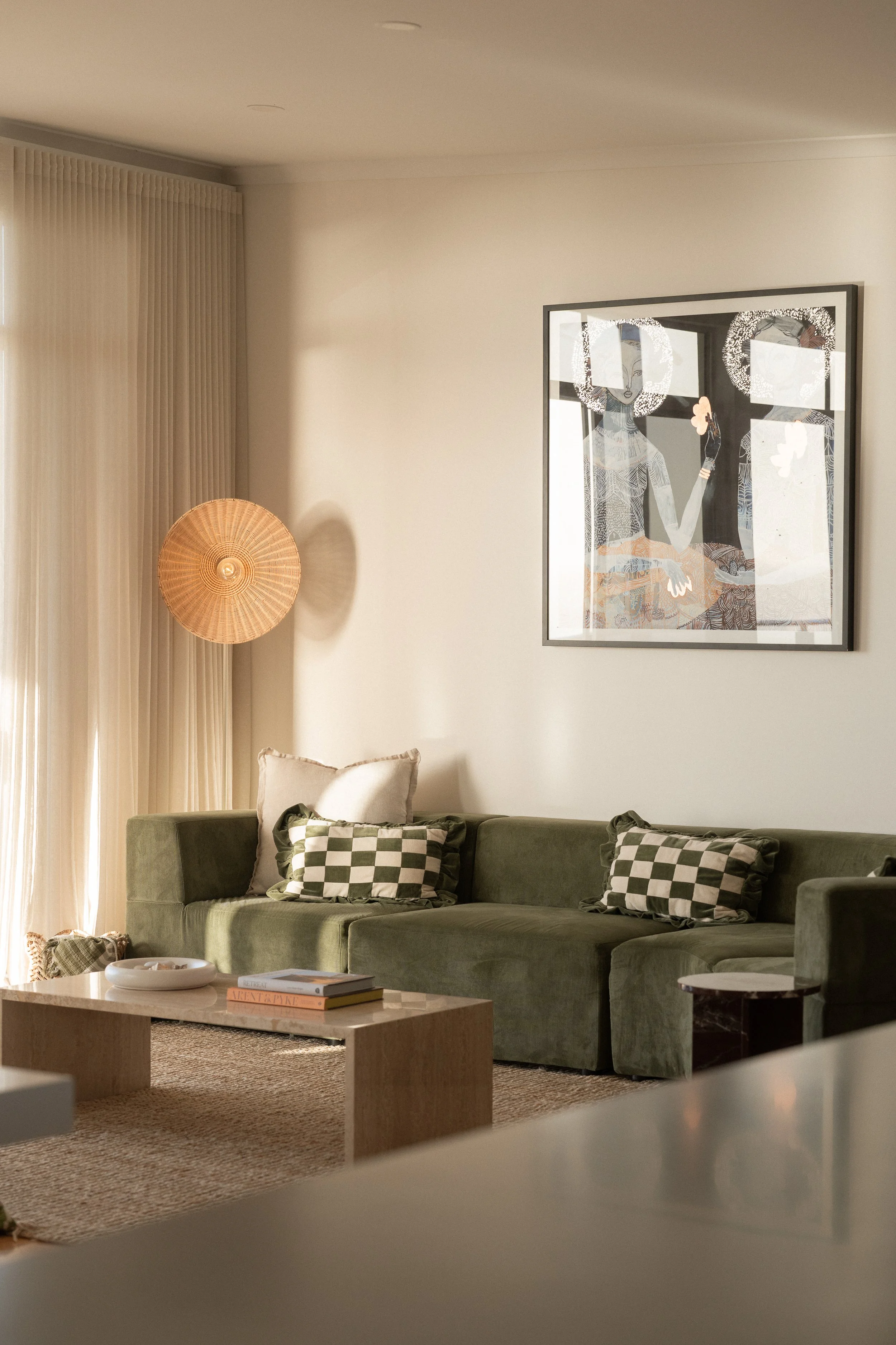 Living room with a green sofa, patterned throw pillows, a coffee table with books, a basket, a wall art piece, a lamp, and curtains.