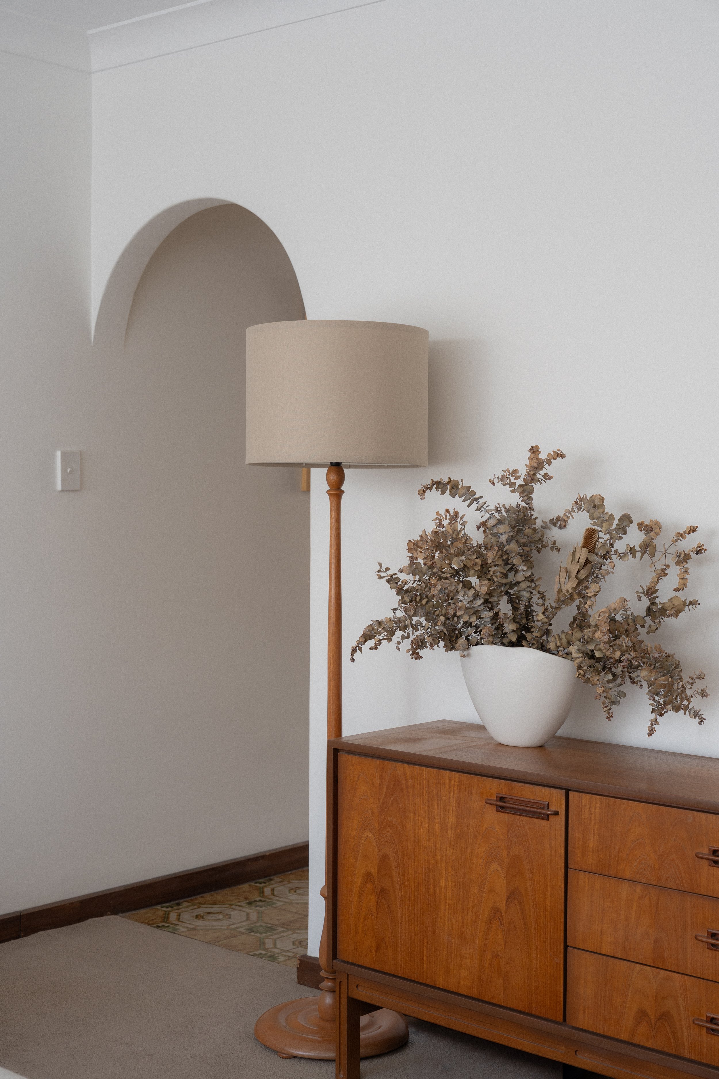 A wooden sideboard with a large white ceramic vase filled with dried eucalyptus branches, next to a beige standing lamp in a room with minimal decor.