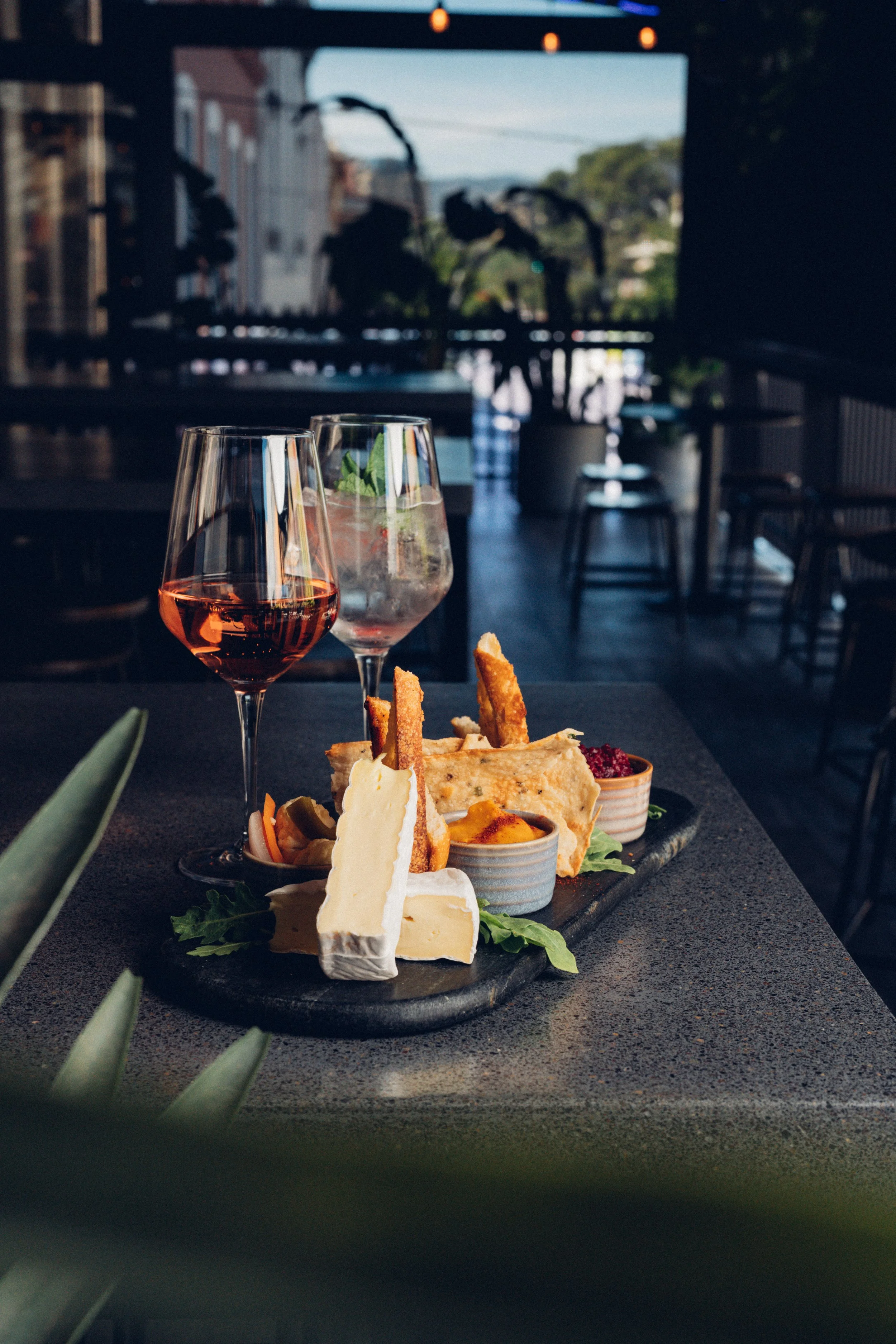 A slate serving platter with cheeses, breadsticks, crackers, and dips on a dark table in a restaurant with outdoor seating. Two glasses of rosé wine are in the foreground, and blurred background of tables and chairs with some greenery and buildings o