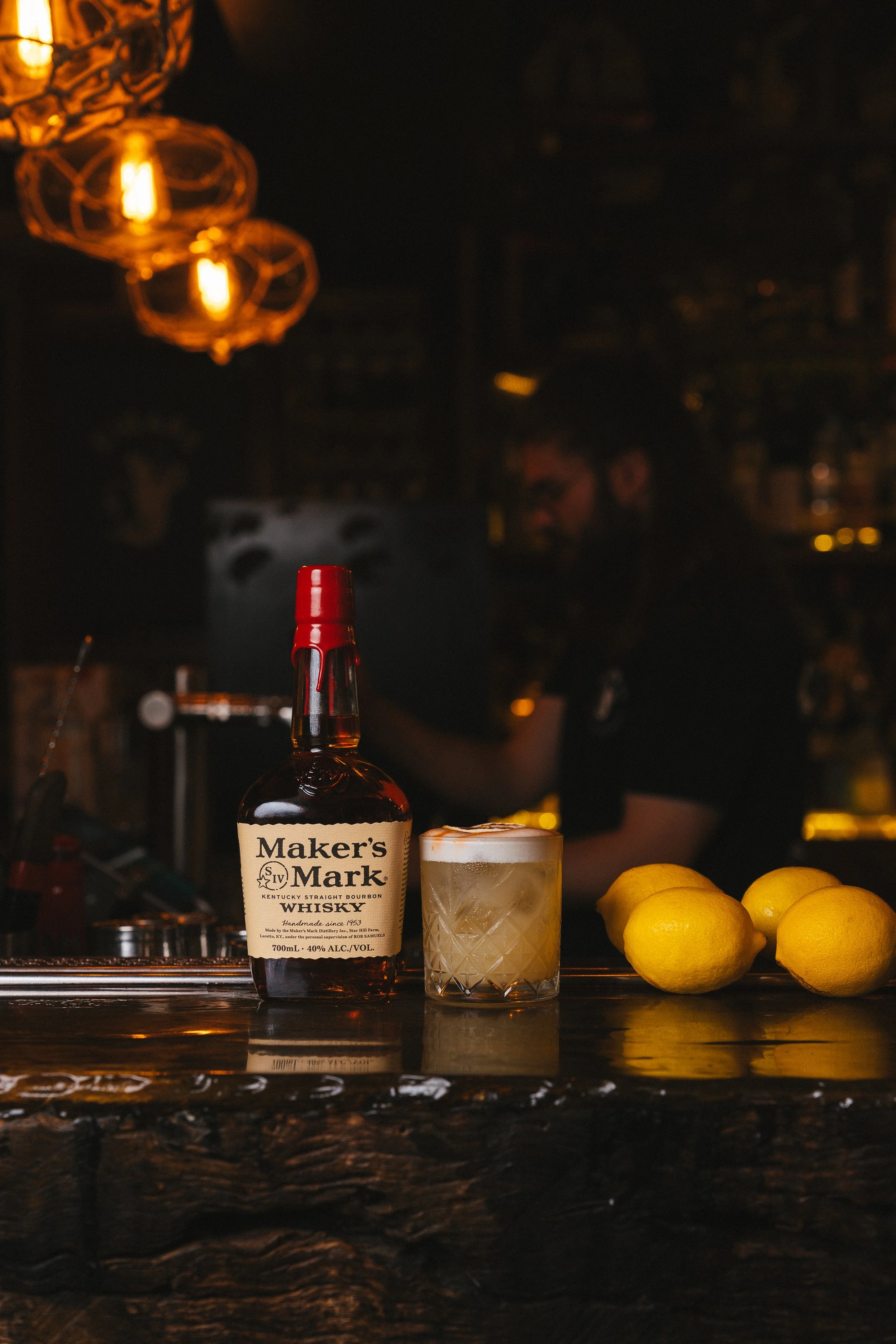 A bar scene with a bottle of Maker's Mark bourbon whiskey, a glass filled with a drink, three lemons, and warm orange lighting from hanging lamps.