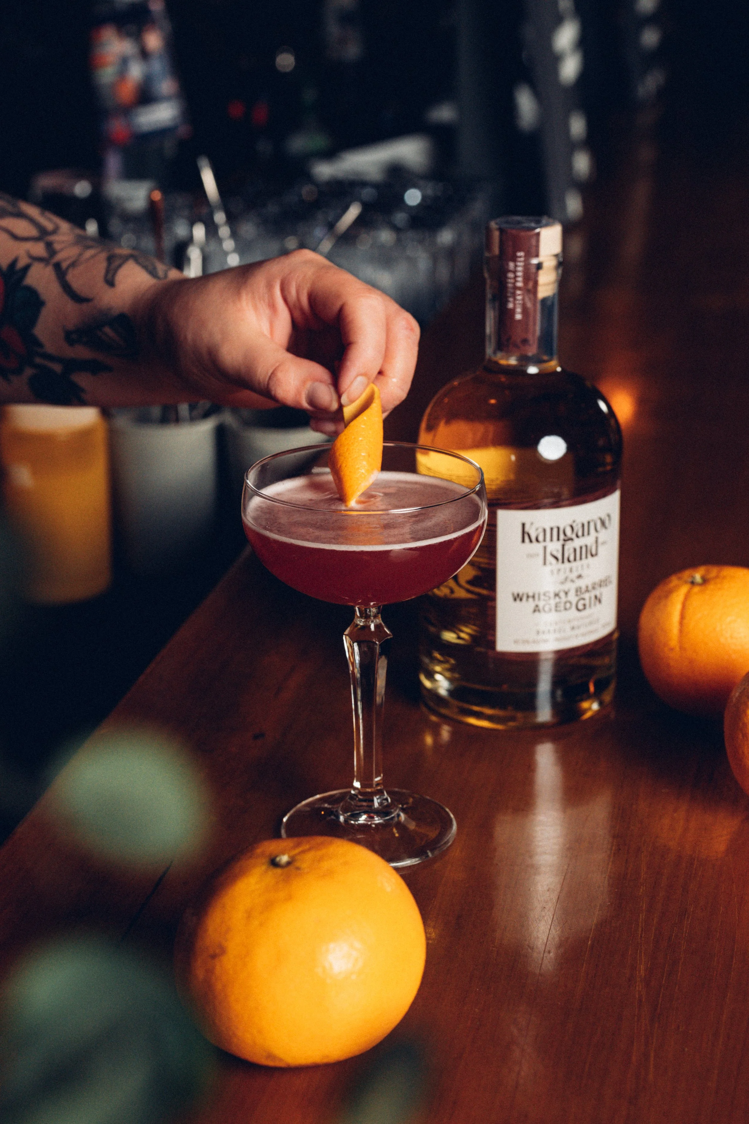 A bar scene with a bottle of Kangaroo Island whisky barrel aged gin, a cocktail glass with a pink drink garnished with a lemon peel, and whole oranges on a wooden surface.