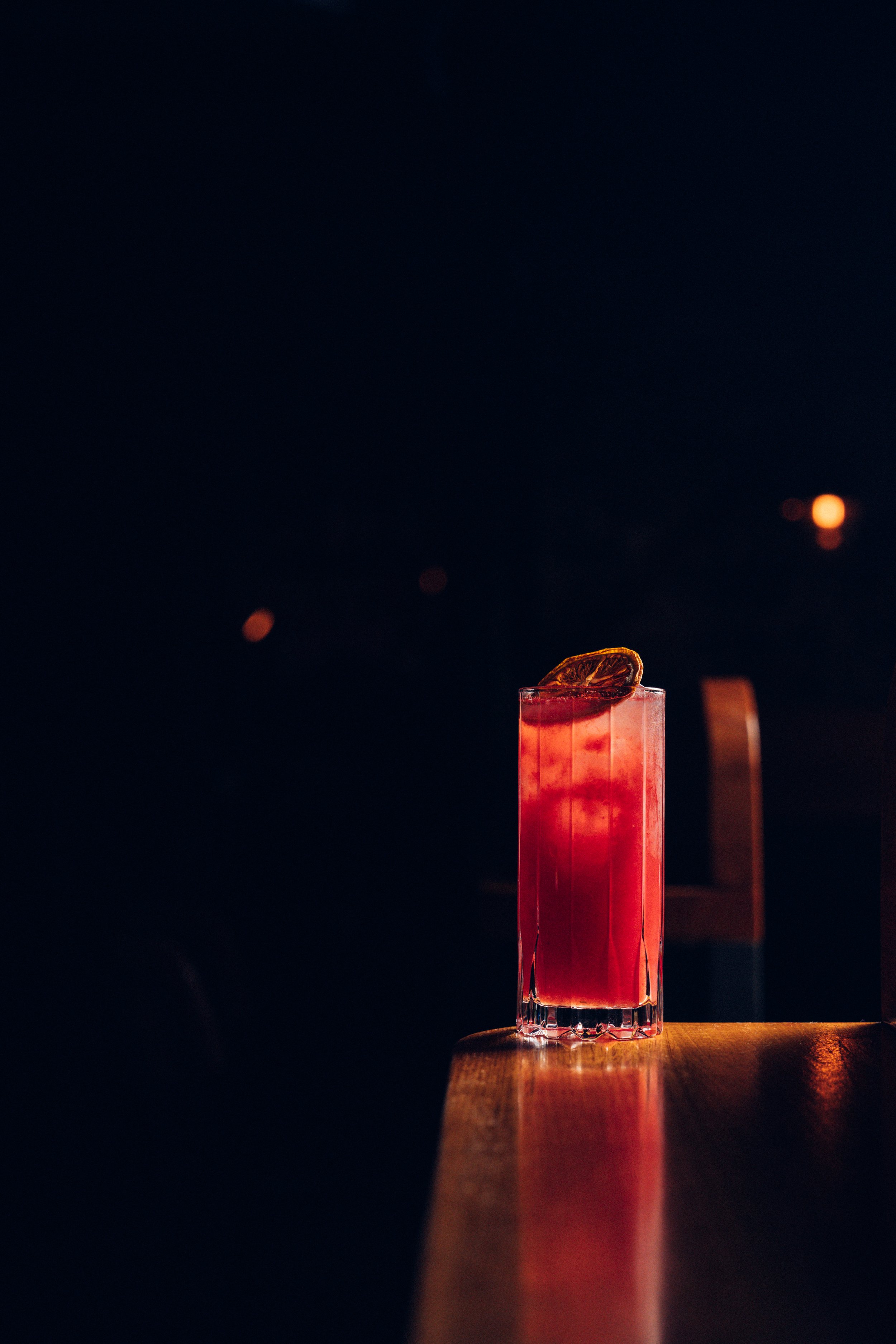 A tall, rectangular glass with a pinkish-red beverage, garnished with a slice of dried citrus, placed on a wooden surface against a dark background with blurred lights.