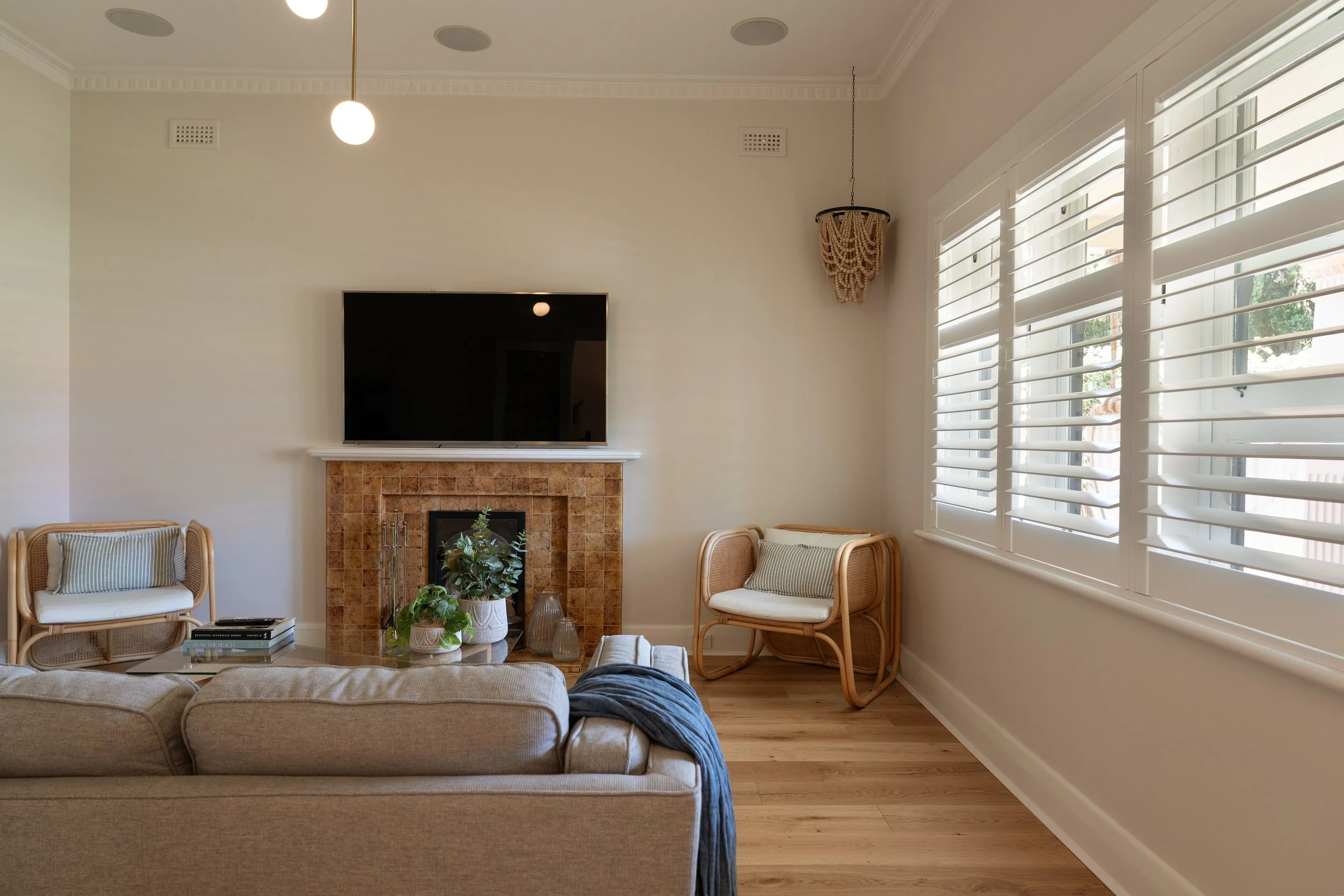 Living room with beige sofa, wood floor, a fireplace with a plant, and a mounted TV above the fireplace. There are two rattan chairs with cushions near the fireplace, a table with books, and large windows with white blinds. A hanging light fixture an