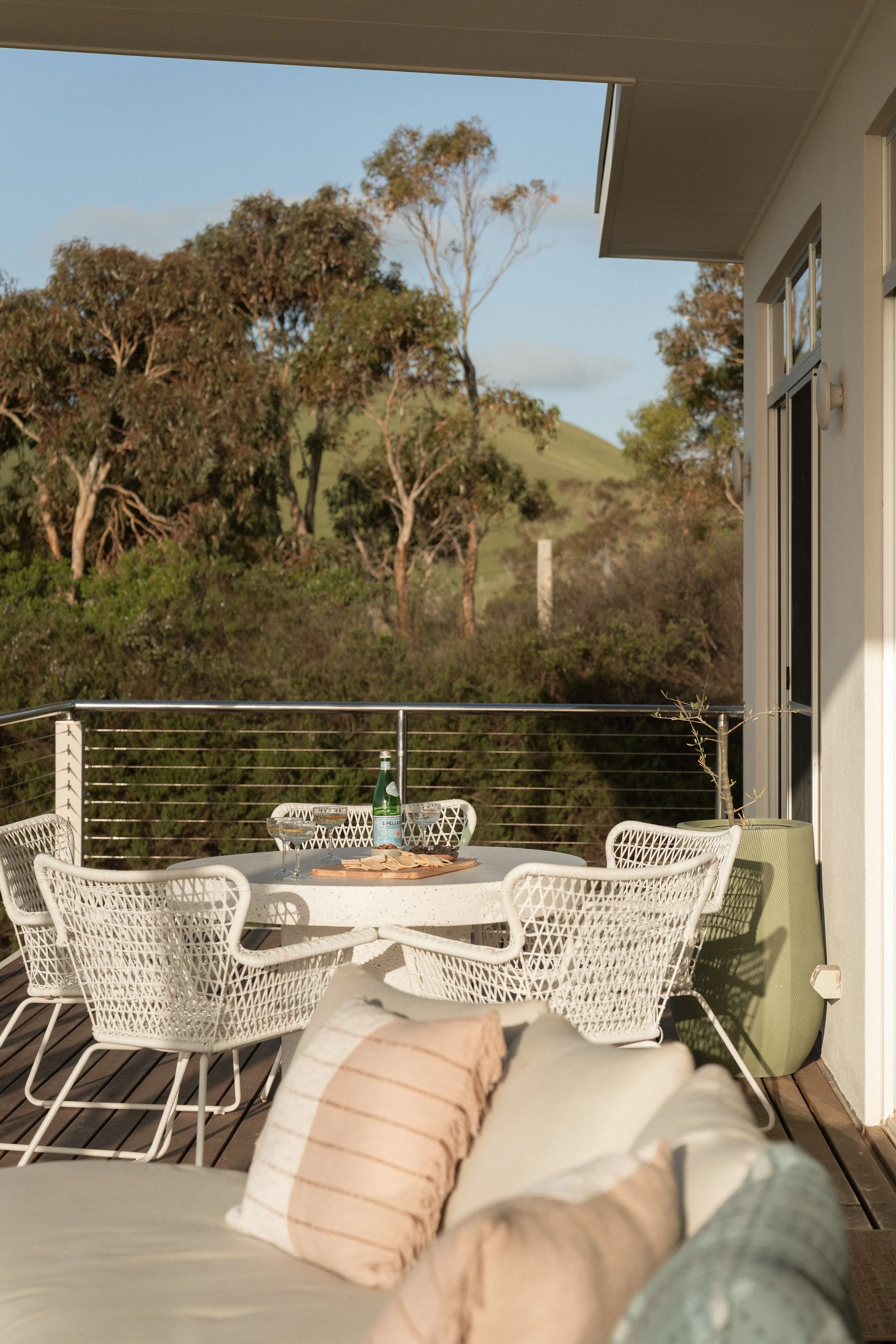 Outdoor balcony with white wicker chairs and a round table set with snacks and drinks, surrounded by a metal railing with a landscape of trees and green hills in the background, and a partial view of a white exterior building with large windows.