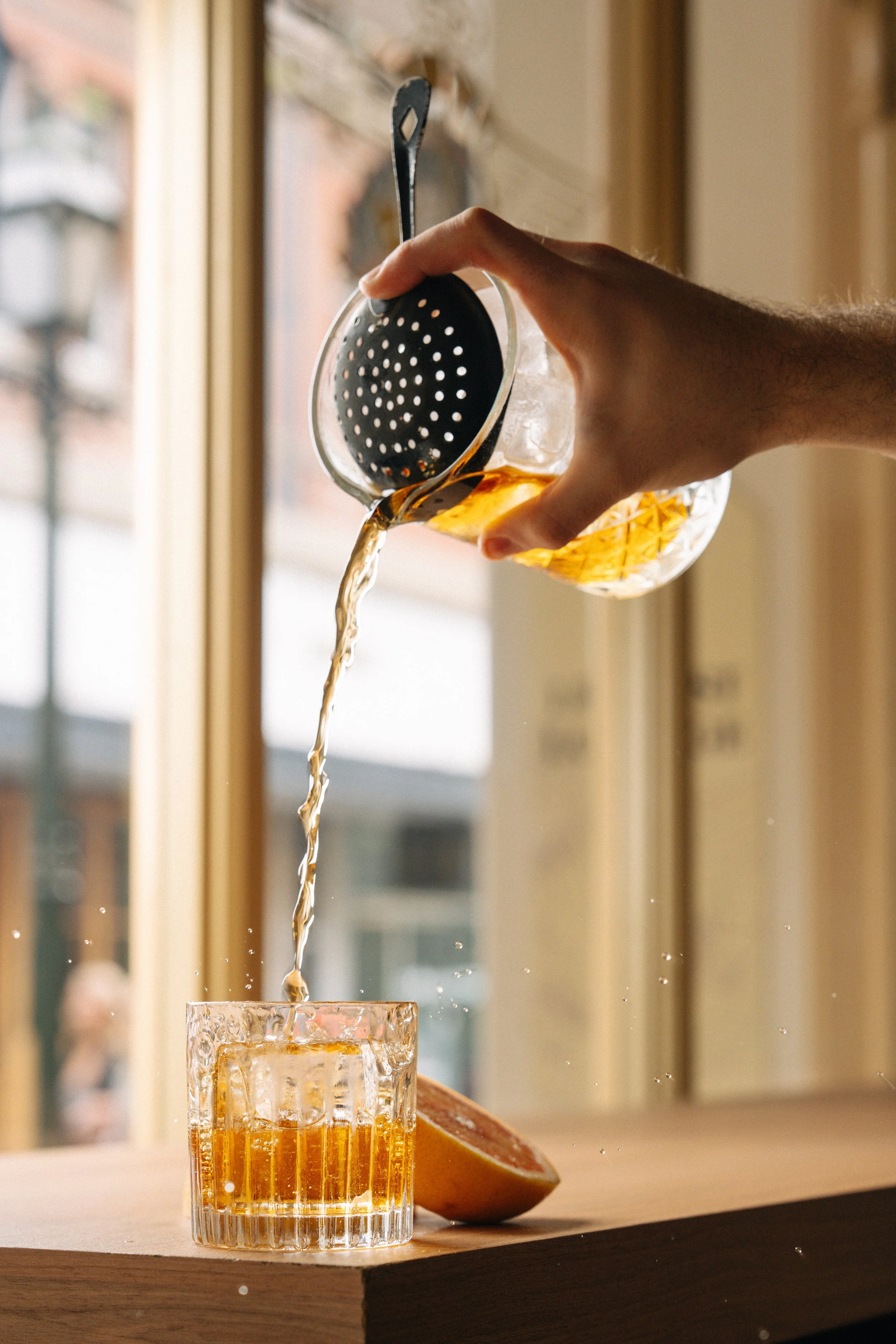 A hand pouring a drink from a glass pitcher into a glass, with a lemon wedge on the table near the glass, set inside a room with large windows.