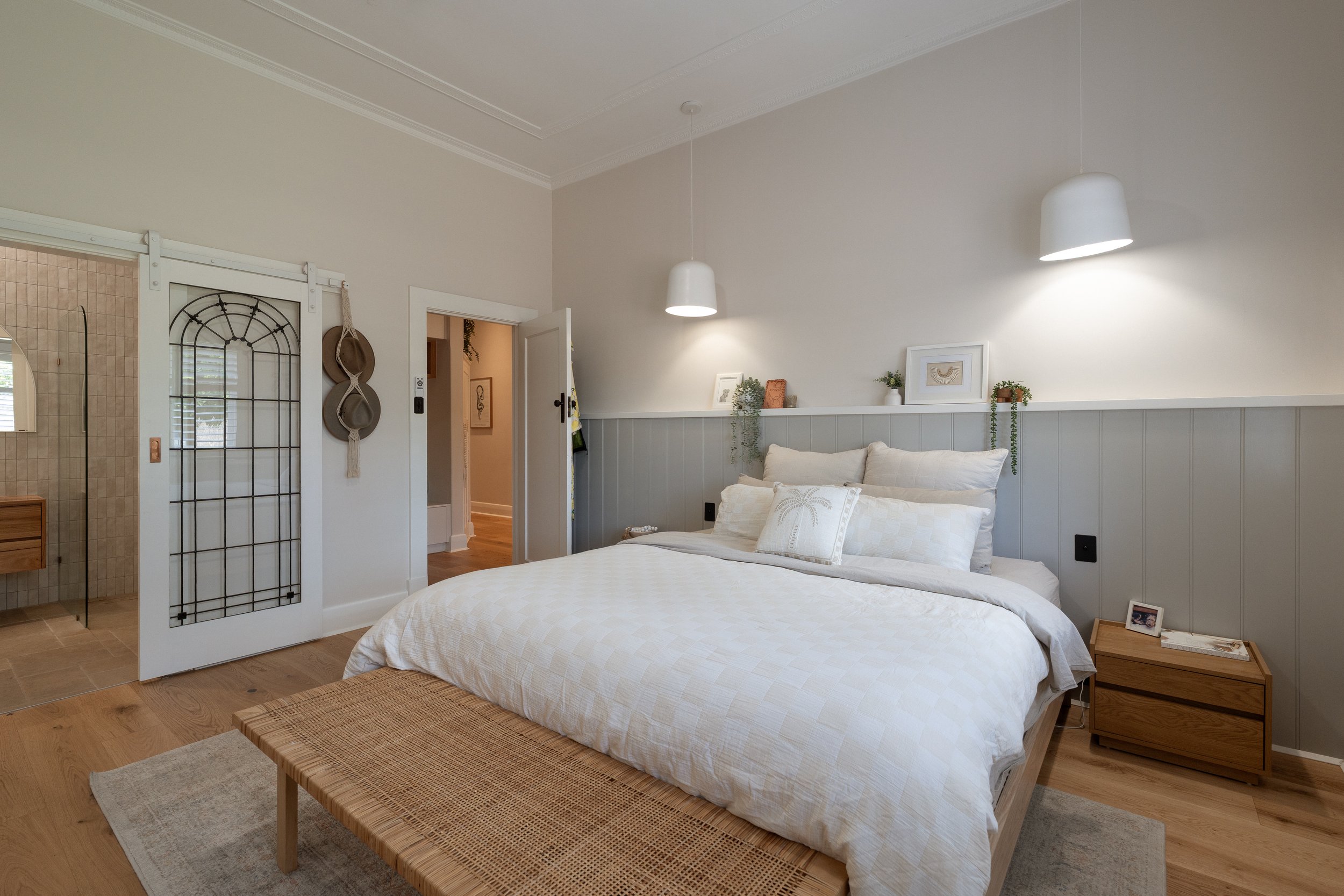 A cozy bedroom featuring a neatly made bed with white bedding and decorative pillows. There are two modern white pendant lights hanging from the ceiling, a light-colored wooden nightstand with framed pictures and books, and a tufted headboard. The wa