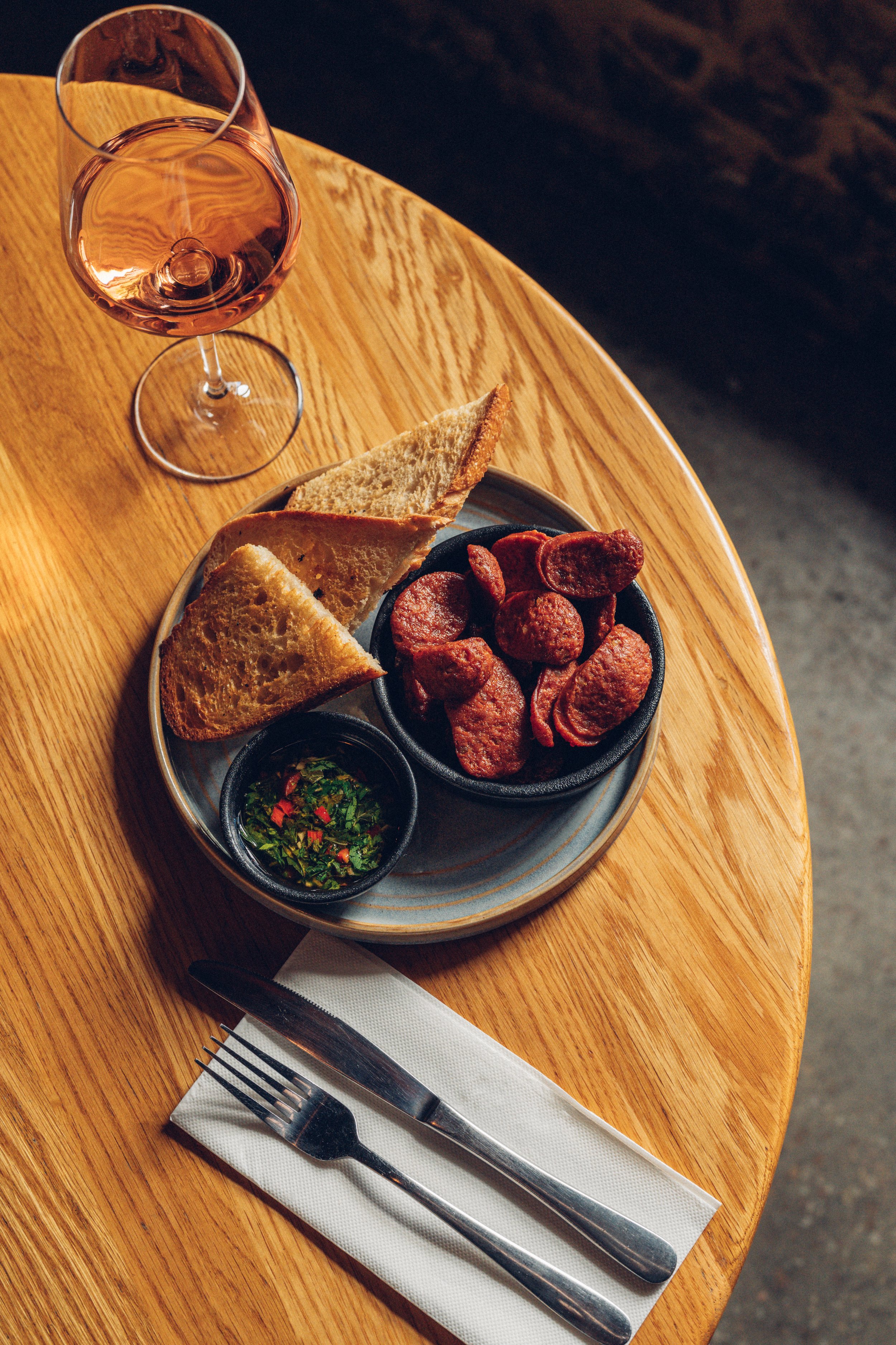 A wine glass filled with pink rosé wine, a plate with slices of bread and a bowl of sliced cured meat, with a small dish of chopped herbs or salsa, on a round wooden table set for dining.