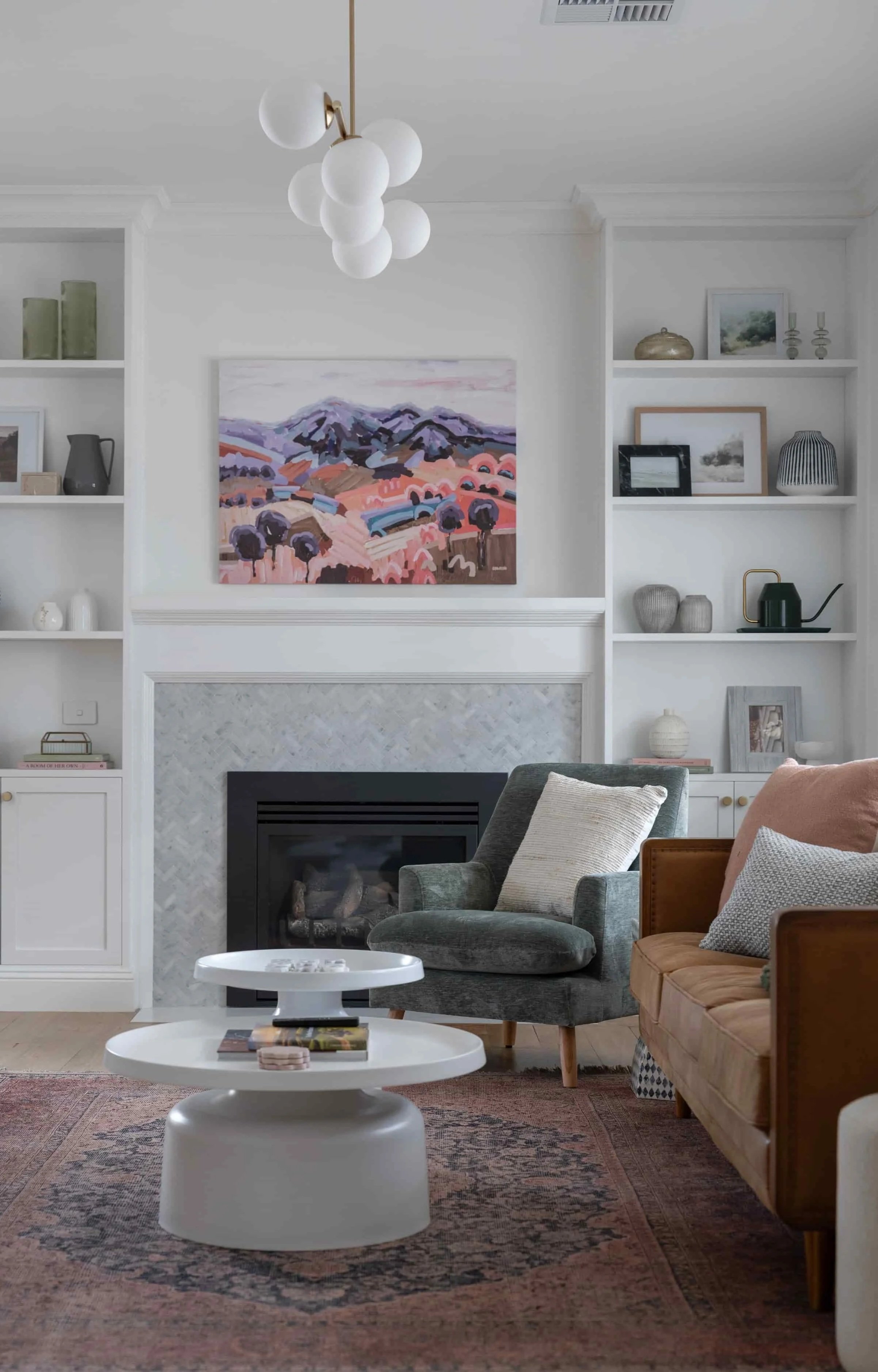 Living room with a fireplace, built-in bookshelves, and a colorful landscape painting above the fireplace. There are two armchairs, one green, one tan, with pillows, in front of a rug, and a white round coffee table with books and objects.