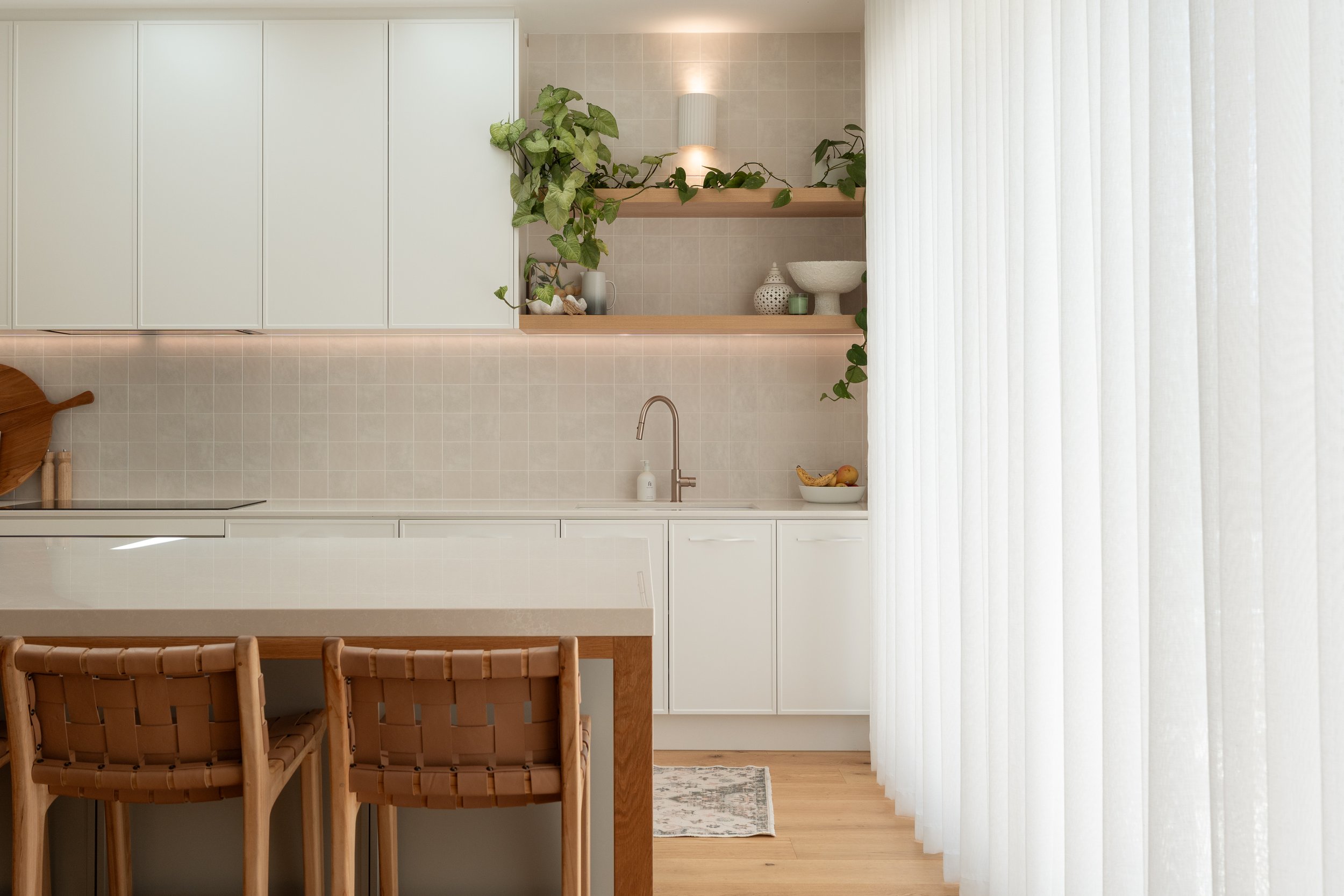 Bright, minimalist kitchen with white cabinets, a wooden table with woven chairs, plants on open shelves, and sheer vertical blinds covering a large window.