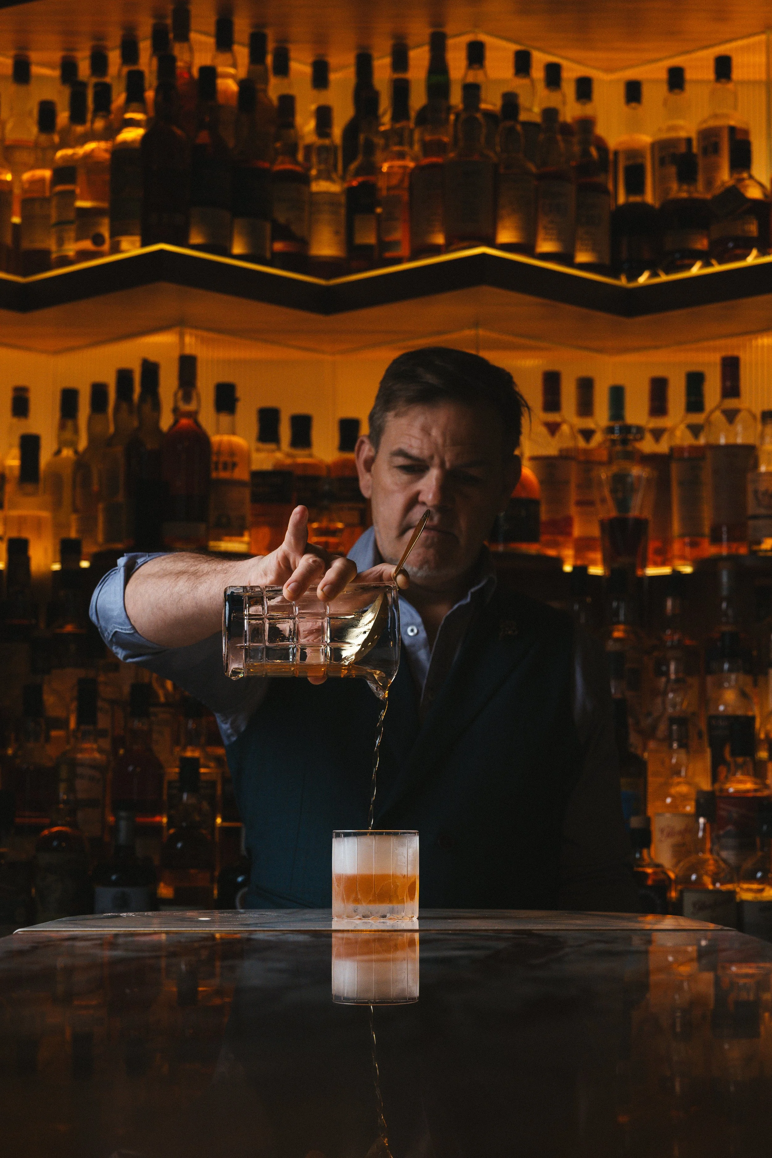 A bartender pouring a drink into a glass at a bar, with shelves of bottles behind him.