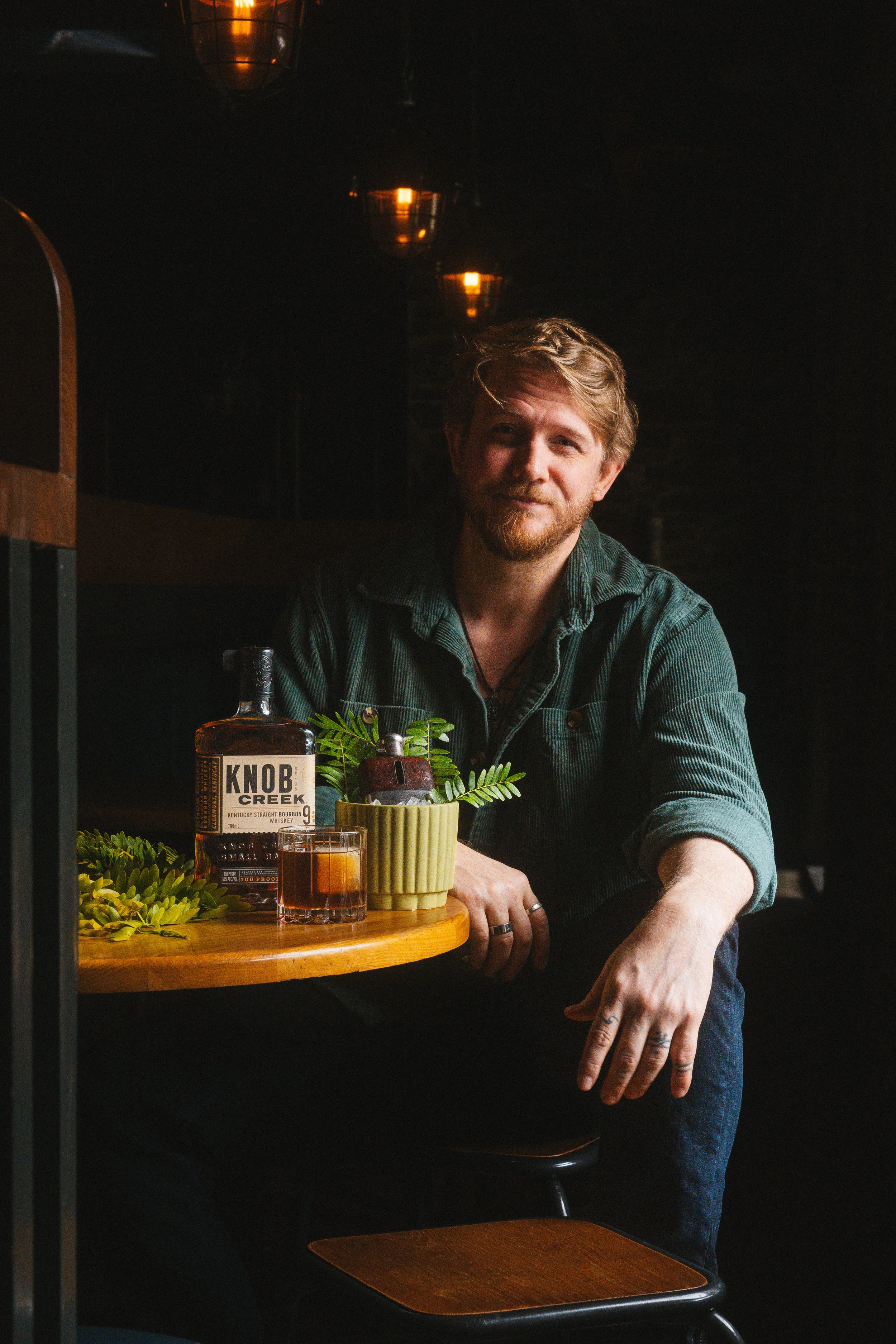 A man with light brown hair and a beard sitting at a restaurant table with a dark background. The table has a bottle of Knob Creek bourbon, a glass of drink, and some green plants. The man is wearing a dark green or teal shirt and has a slight smile,