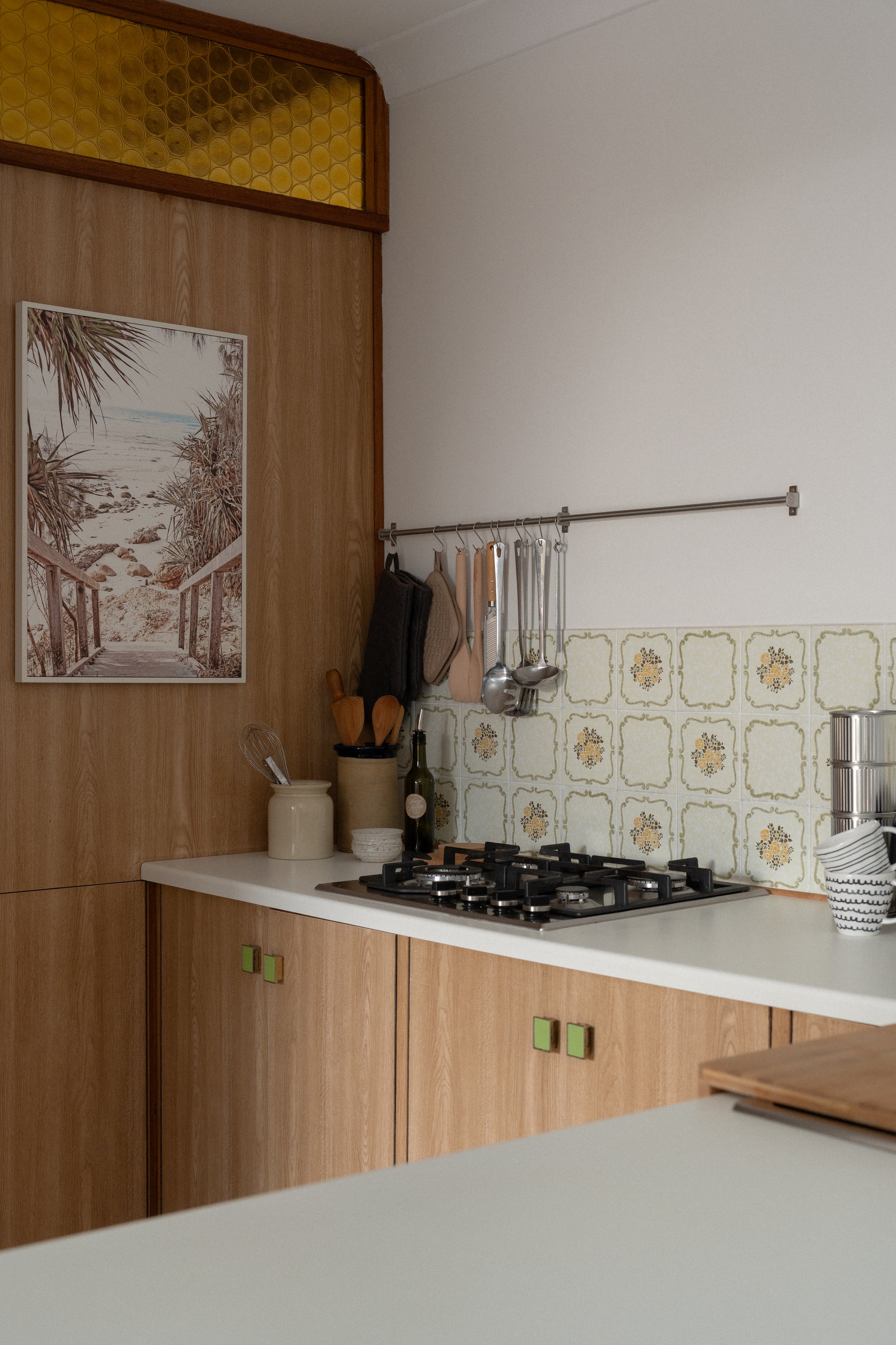A kitchen with wood cabinets, a white countertop, and a white tiled backsplash with yellow floral patterns. Above the counter, a metal rod holds various kitchen utensils and bags. There is a picture of a beach scene on the wall, and a small section o