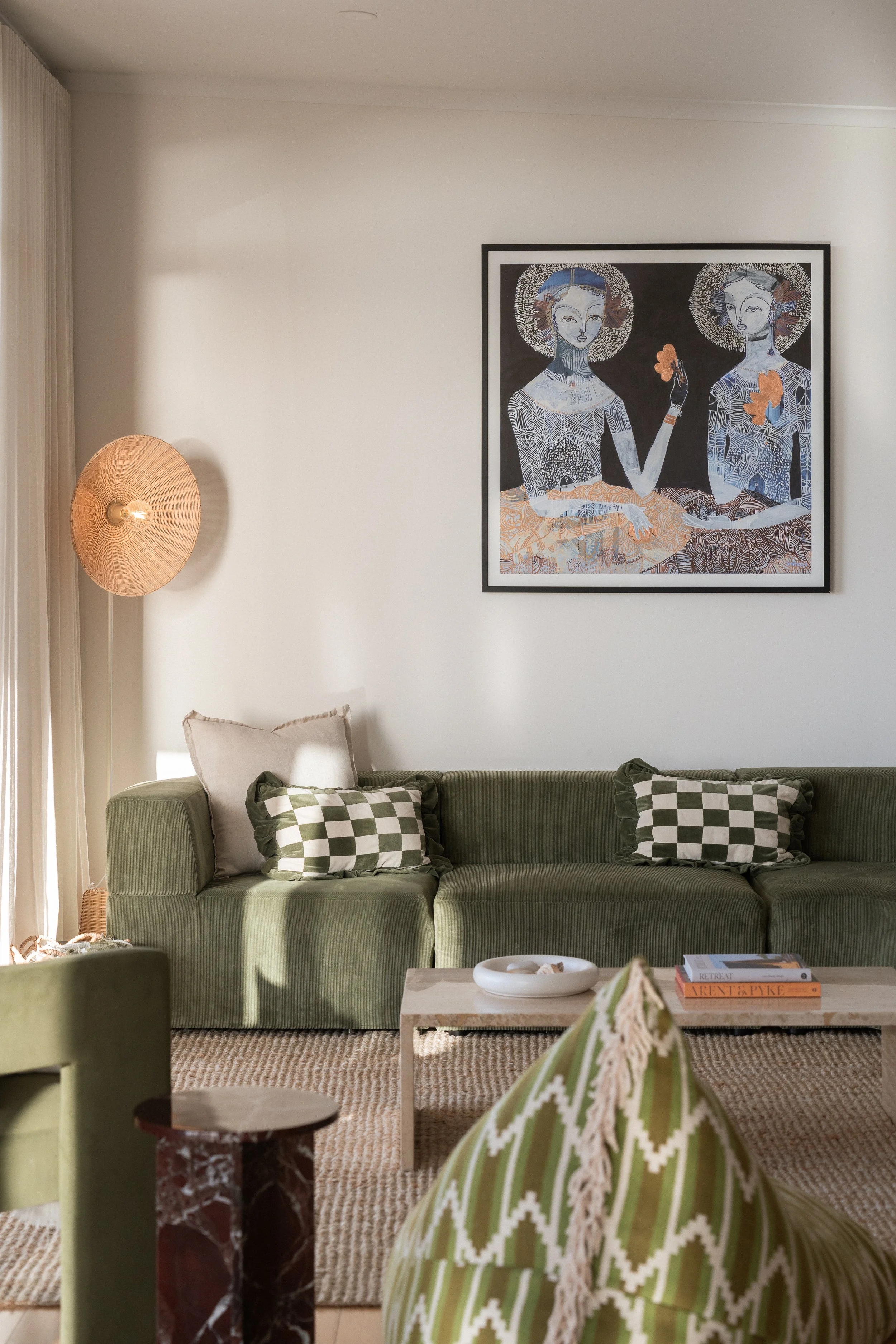 Living room with a green sofa, checkered pillows, a coffee table with books and a bowl, a textured rug, a wicker wall lamp, and artwork of two women hanging on the white wall.