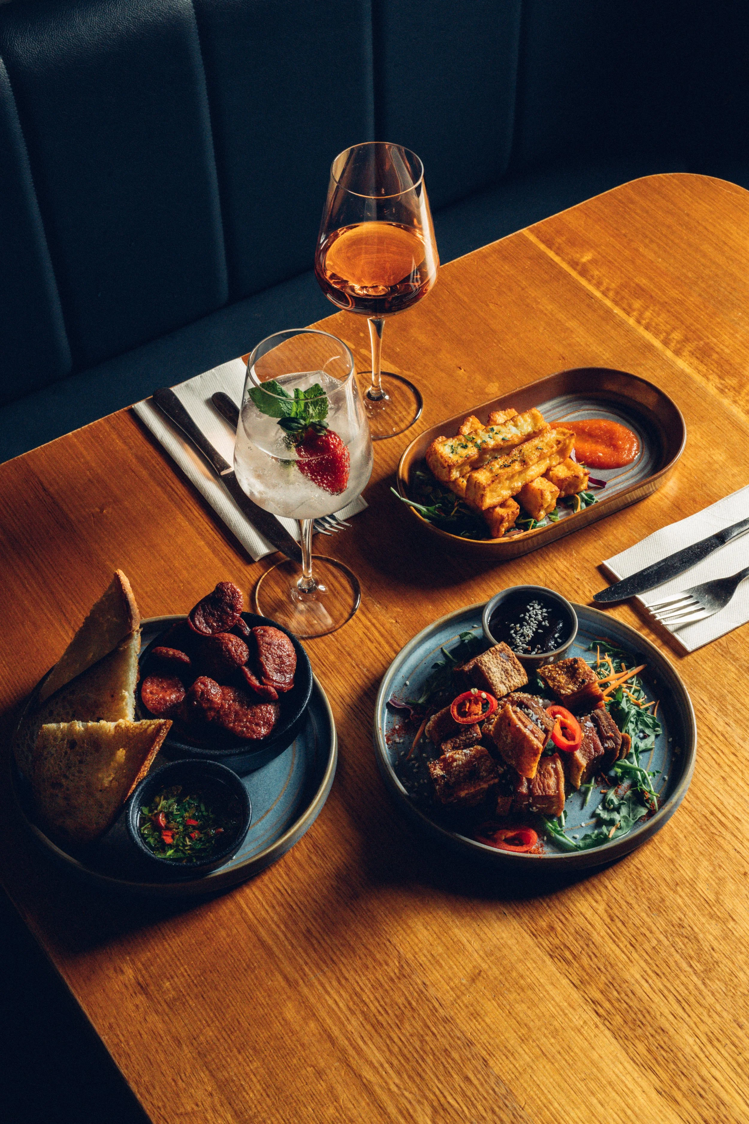 Two glasses of drinks, one with red wine, one with a strawberry mint cocktail, along with various dishes including fried tofu, sliced meats, spring roll, and Asian-style salads on a wooden table in a restaurant setting.