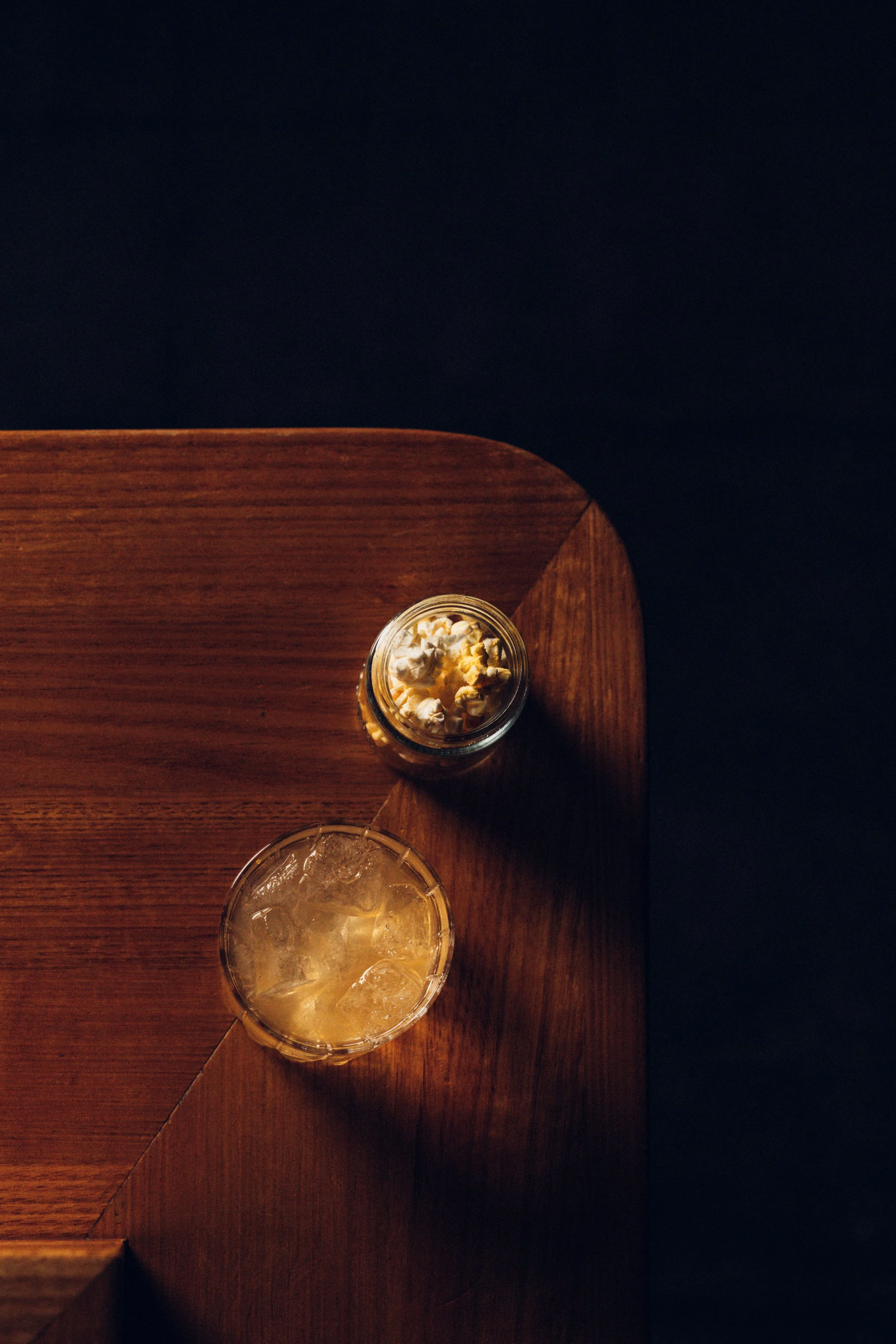 Top view of a wooden table with two glasses of beverages, one containing ice and a light-colored drink, and another with popcorn inside a small jar, against a dark background.