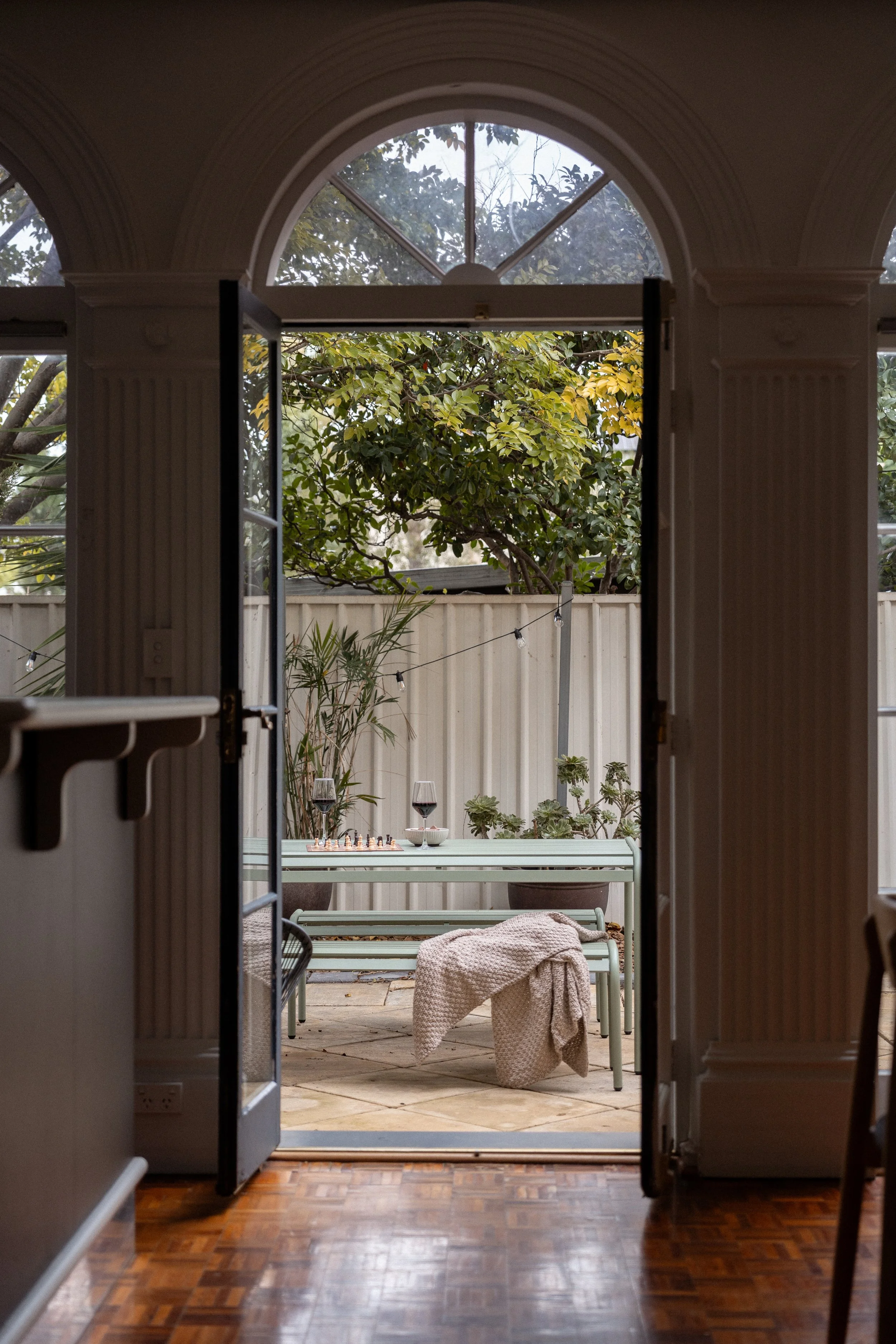 View from inside the house through open double doors to a backyard patio with plants, a fence, a table with two glasses of red wine, a chessboard, and a blanket-covered dress form.
