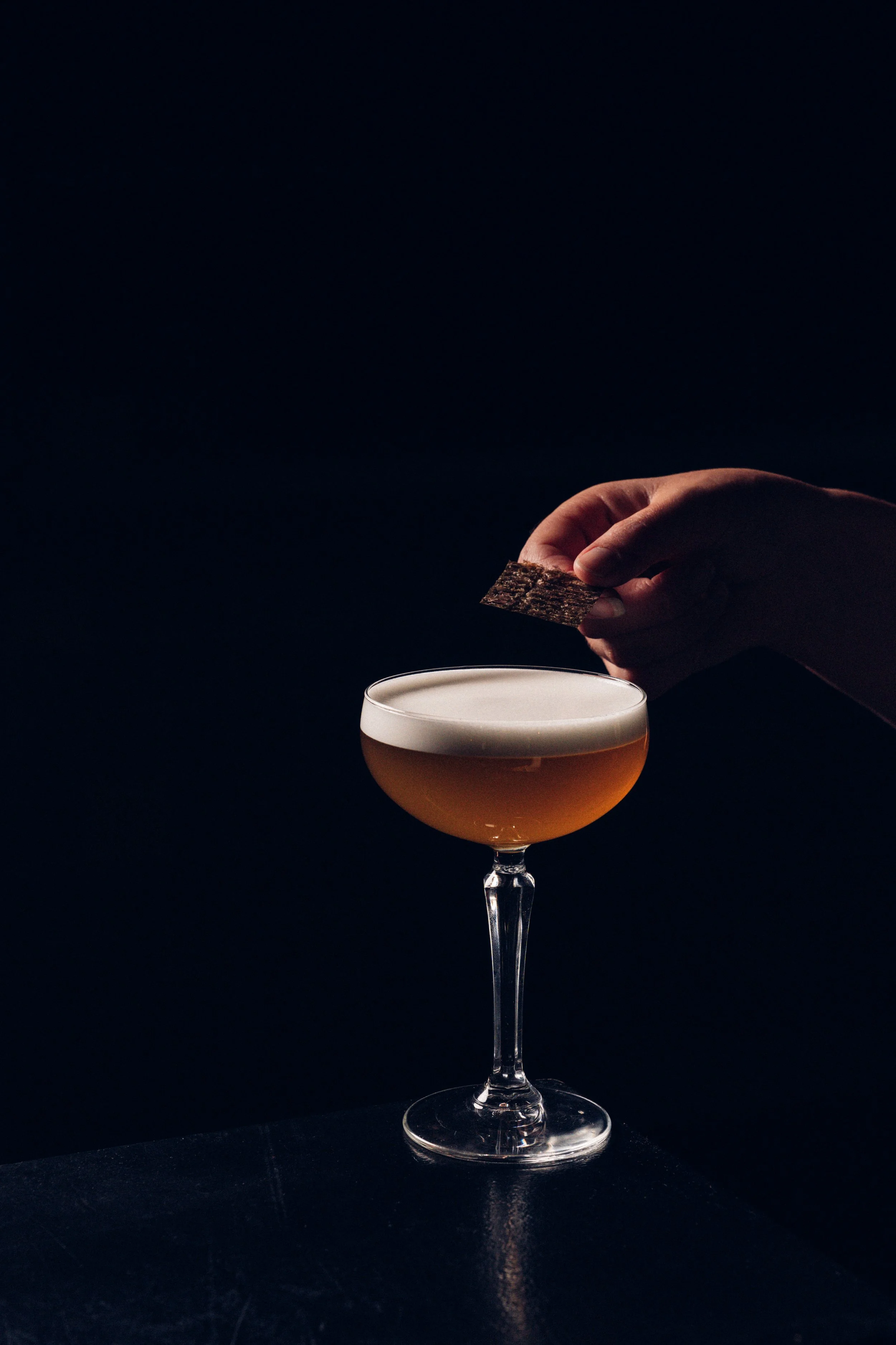 A hand placing a piece of bark-like material into a cocktail glass with a light orange-colored drink, against a dark background.