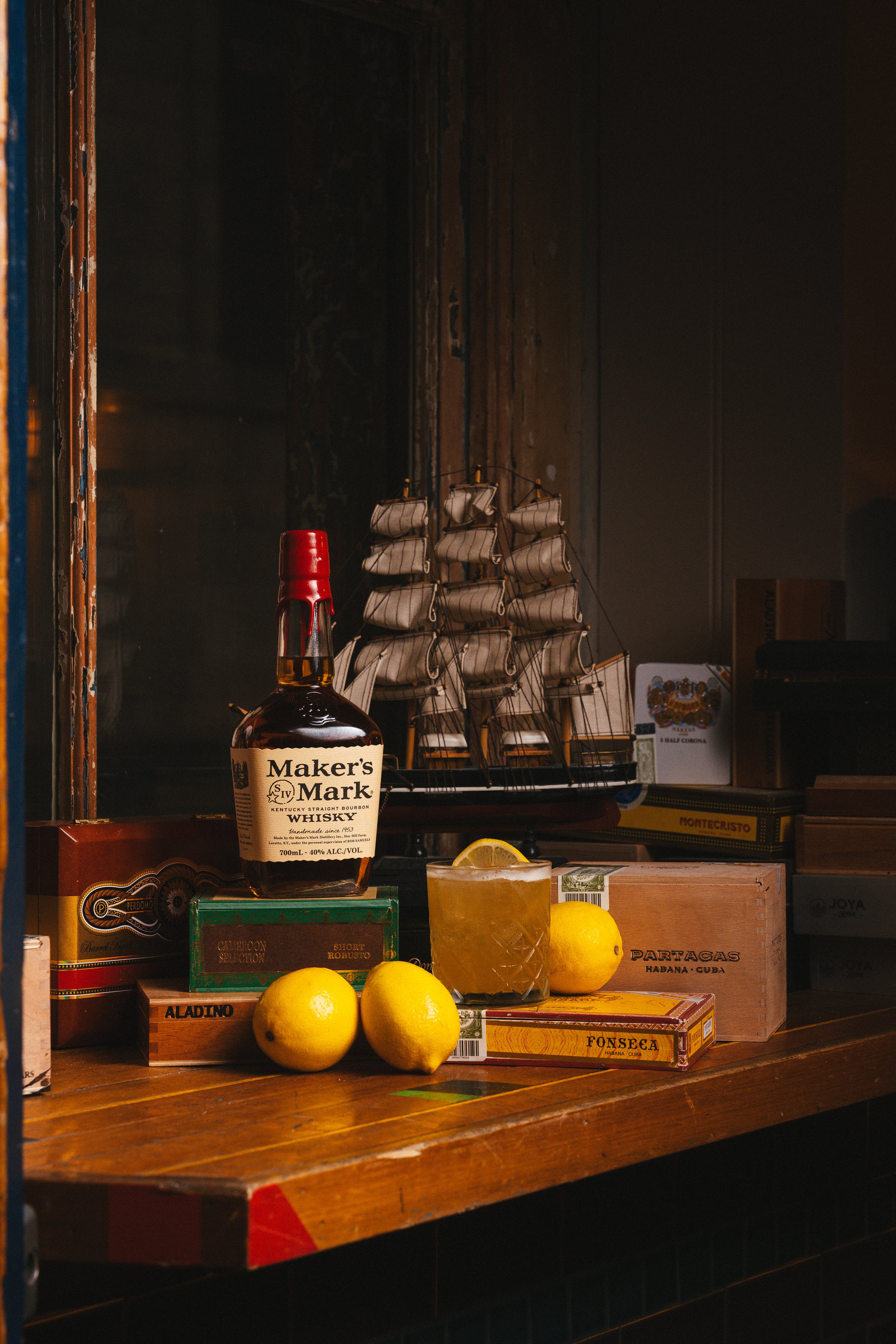 A bottle of Maker's Mark whiskey, three yellow lemons, and a glass of lemonade on a wooden table, with a model ship, lemons, and cigar boxes in the background.