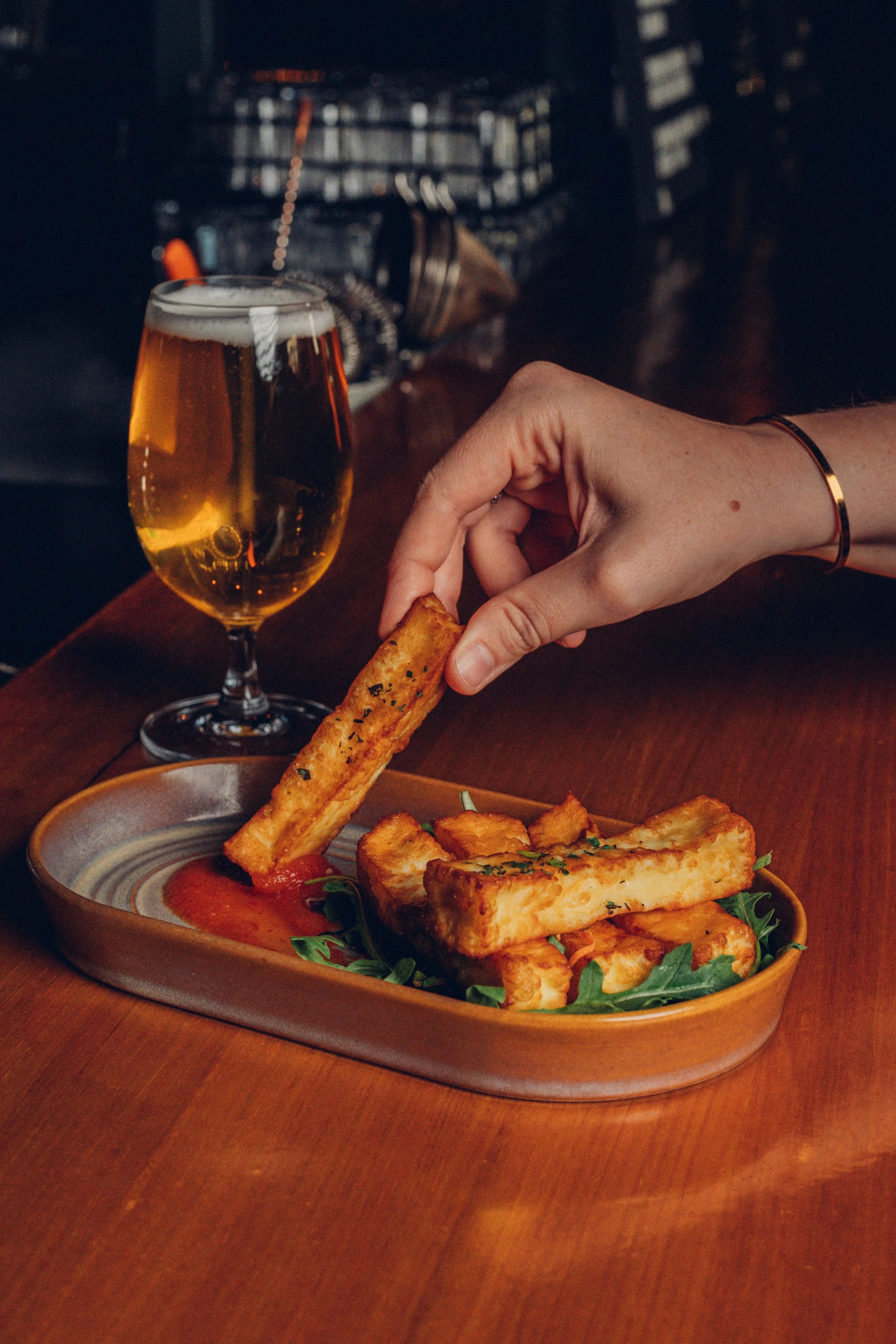 A hand dipping a fried mozzarella stick into marinara sauce on a wooden table, with a glass of beer and a plate of mozzarella sticks in the background.