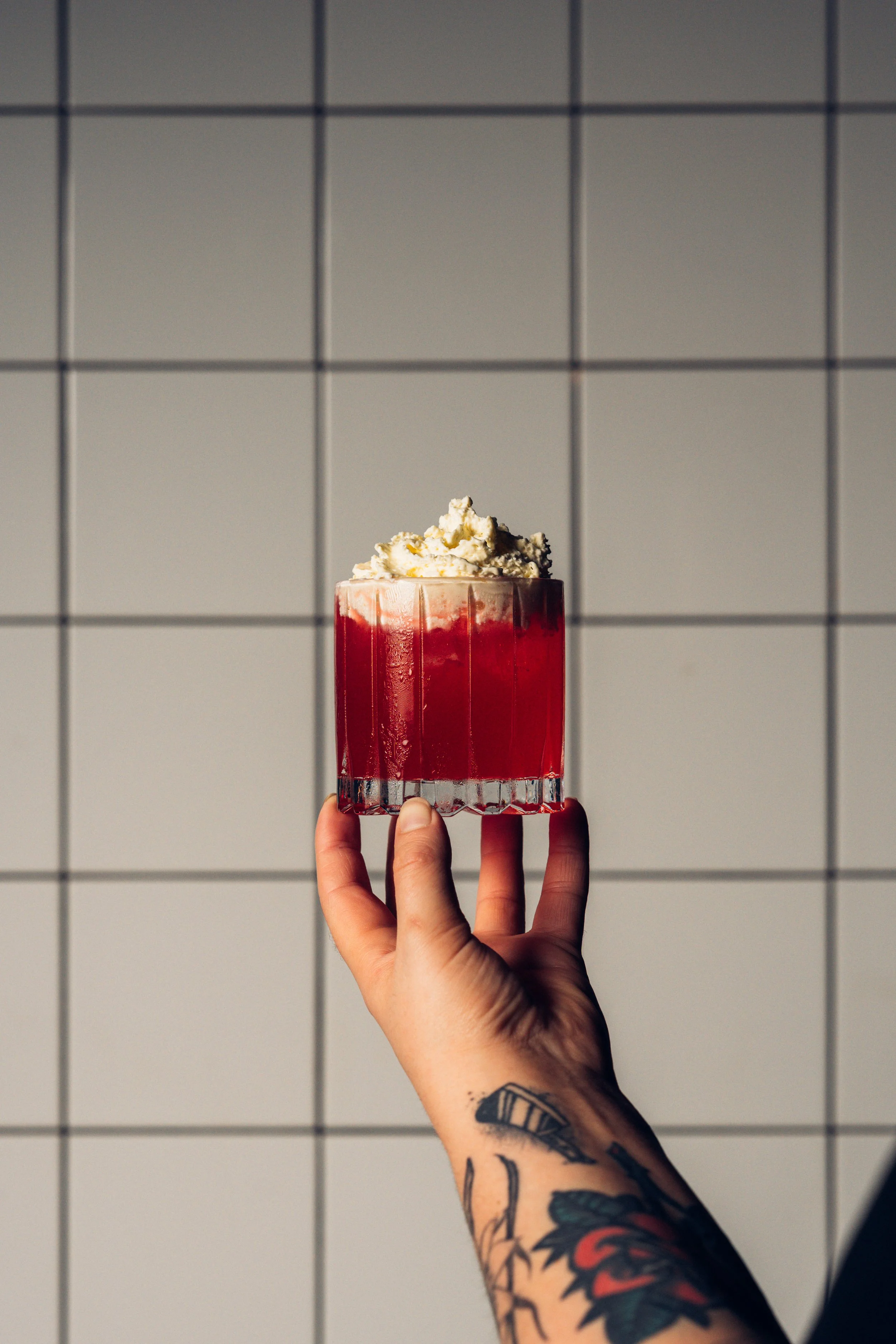A person with tattooed arm holding a glass of red beverage topped with whipped cream against a tiled wall.