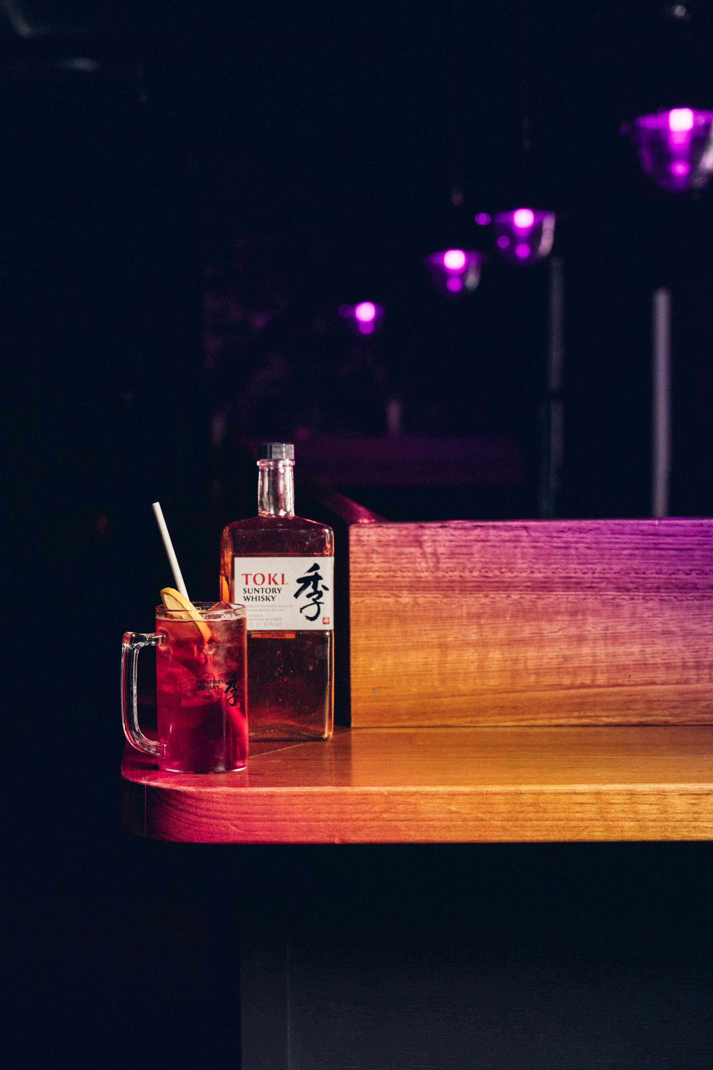A wooden counter with a bottle of Toki whisky, a glass with a red drink, a slice of lemon, and a white straw. In the background, purple-lit glasses are hanging upside down.